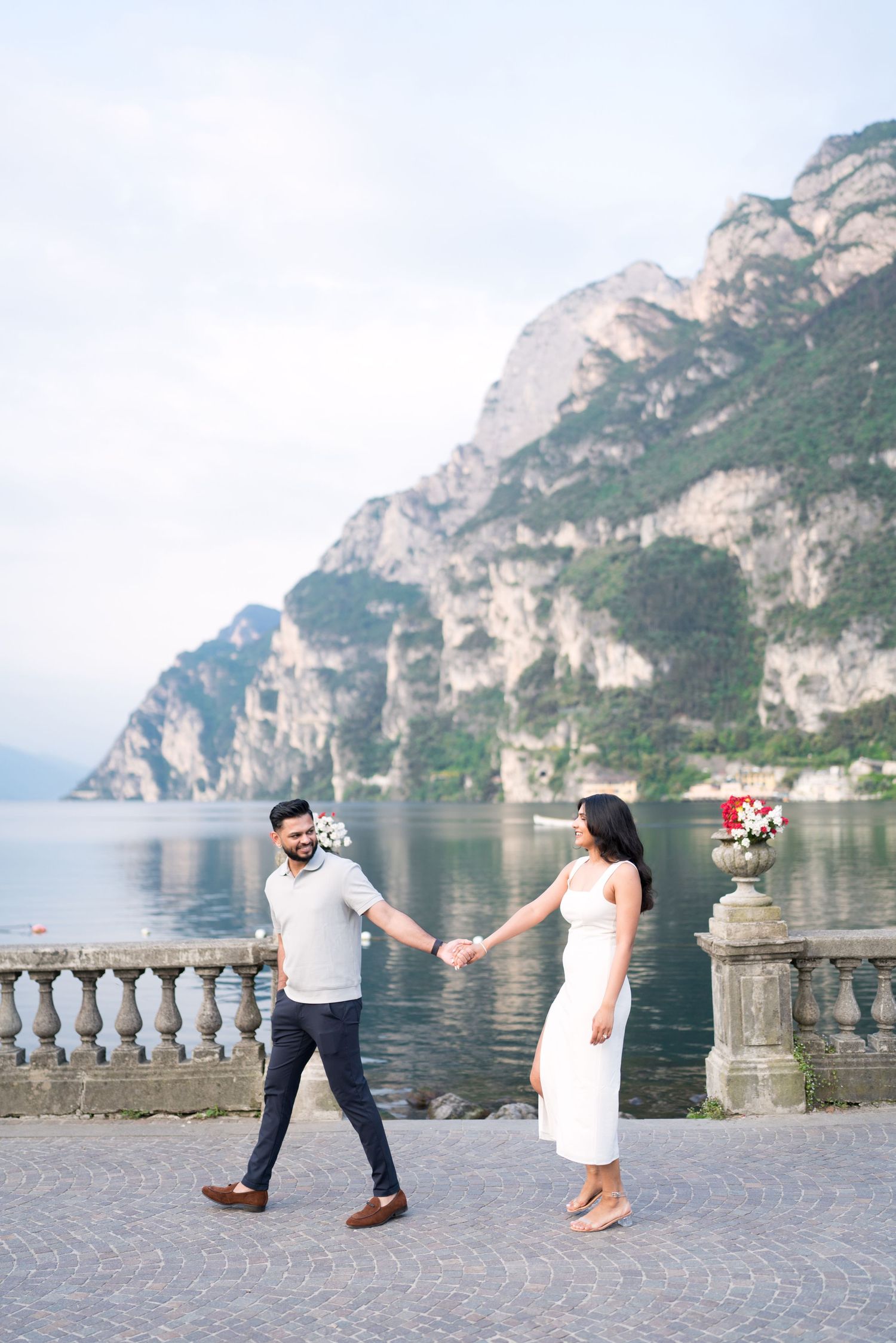 Couple holding hands on lakeside promenade with dramatic mountain cliffs of Lake Garda, Italy in background.