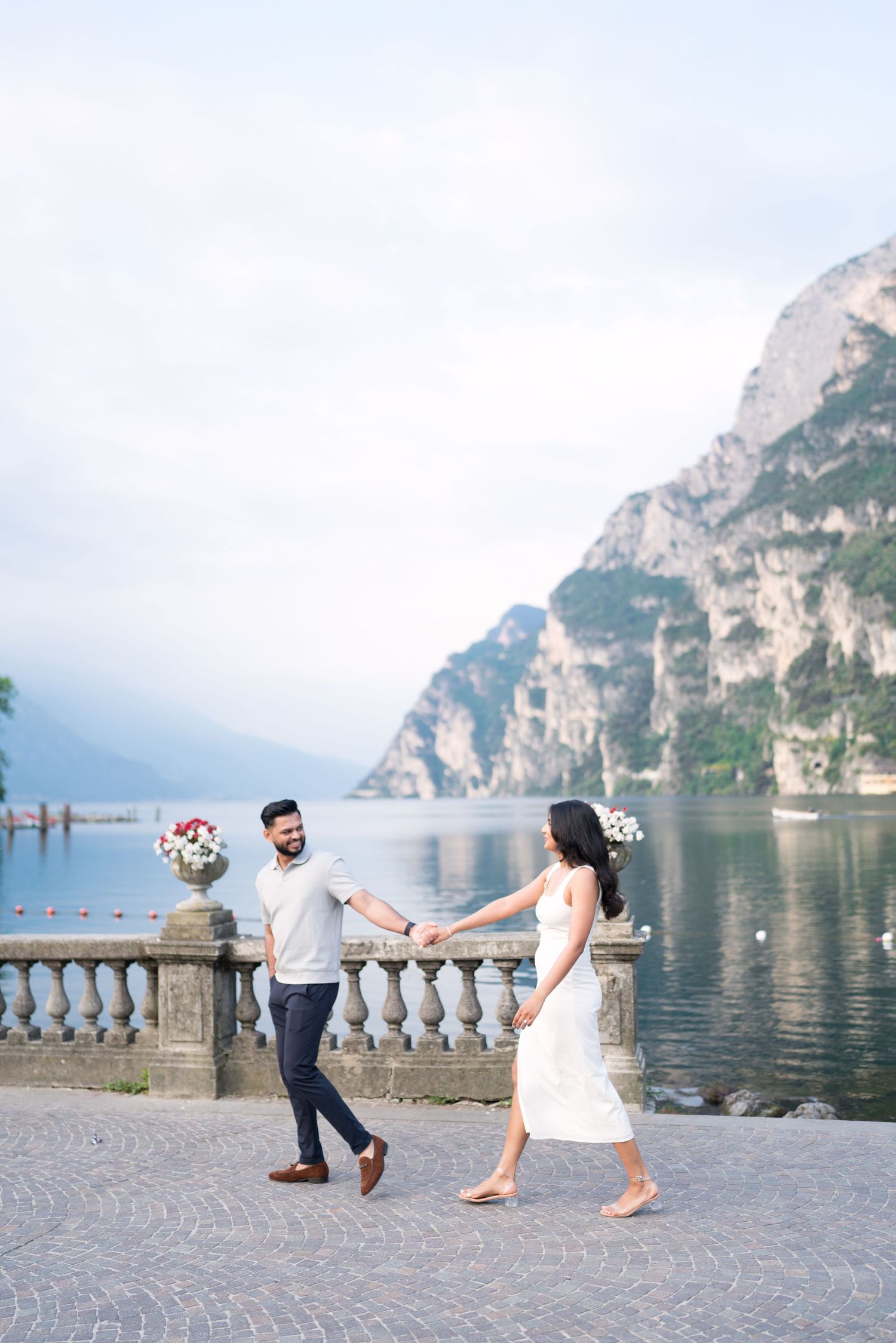 A romantic couple in wedding attire poses playfully by a scenic lakeside balustrade with mountain views in Lake Garda, Italy.