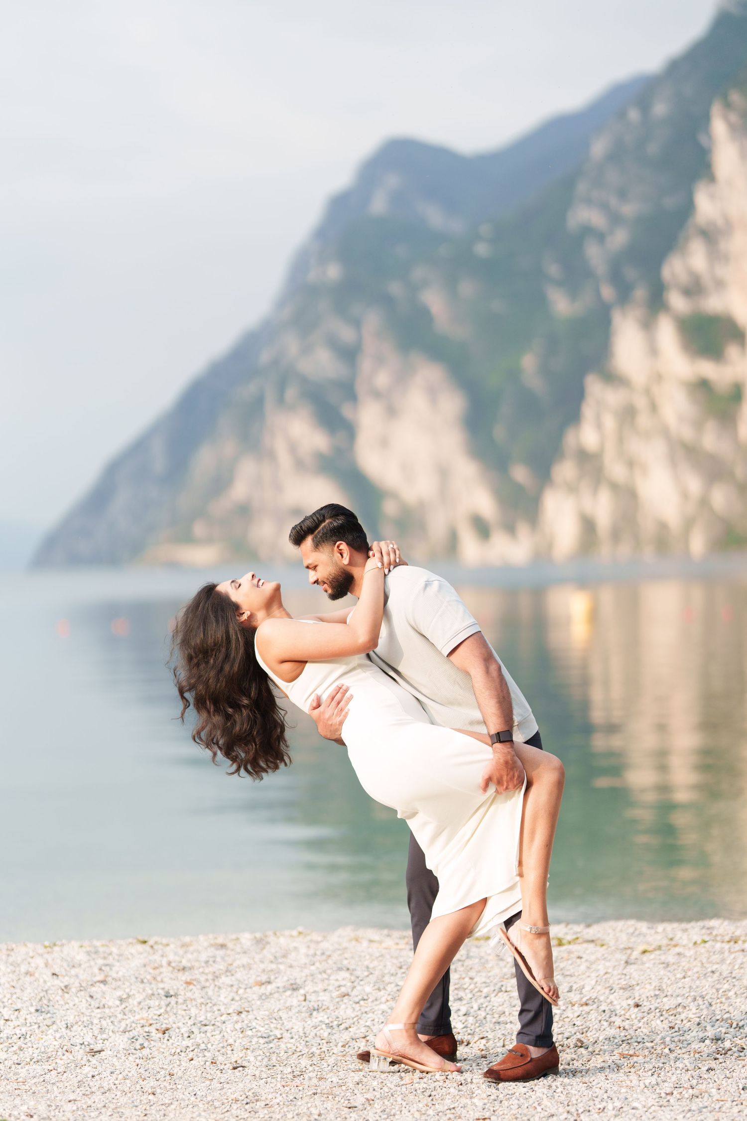 Romantic couple embraces on a pebble beach with dramatic mountain cliffs and turquoise waters in the background.