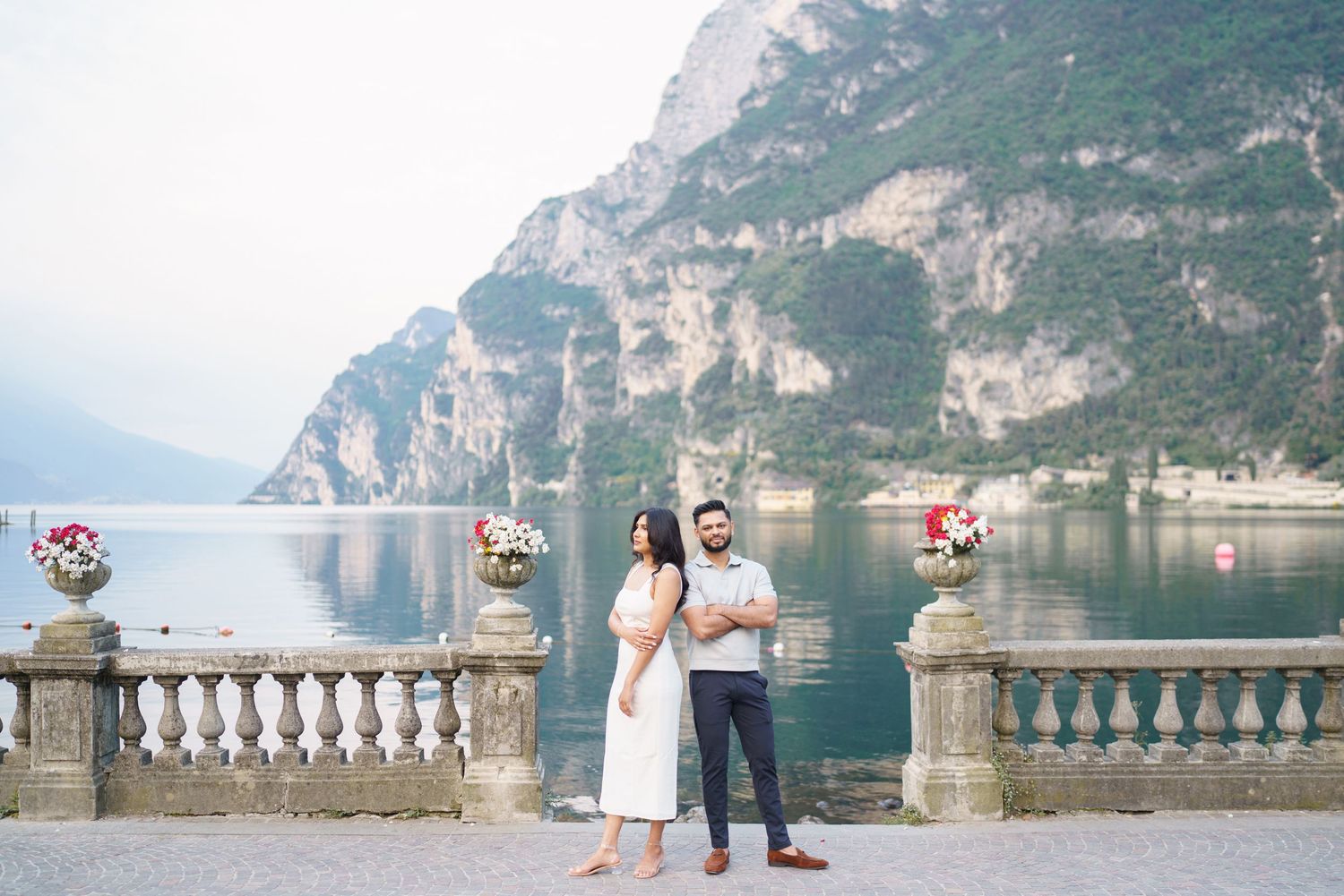 A couple stands on a stone balcony overlooking Lake Como with dramatic mountain cliffs in the background.