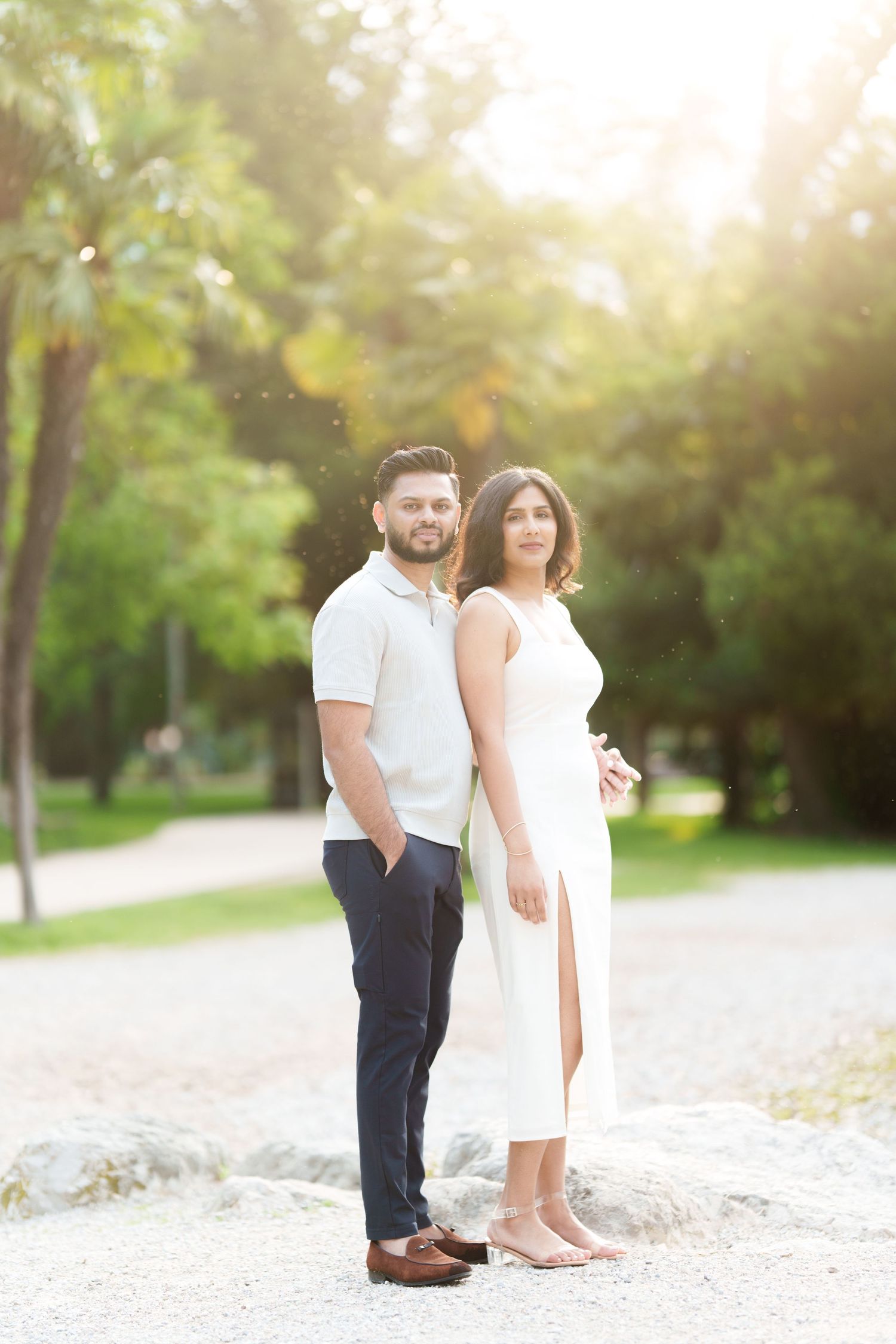 A couple stands together on a sandy beach path during golden hour with sunlit trees in the background.