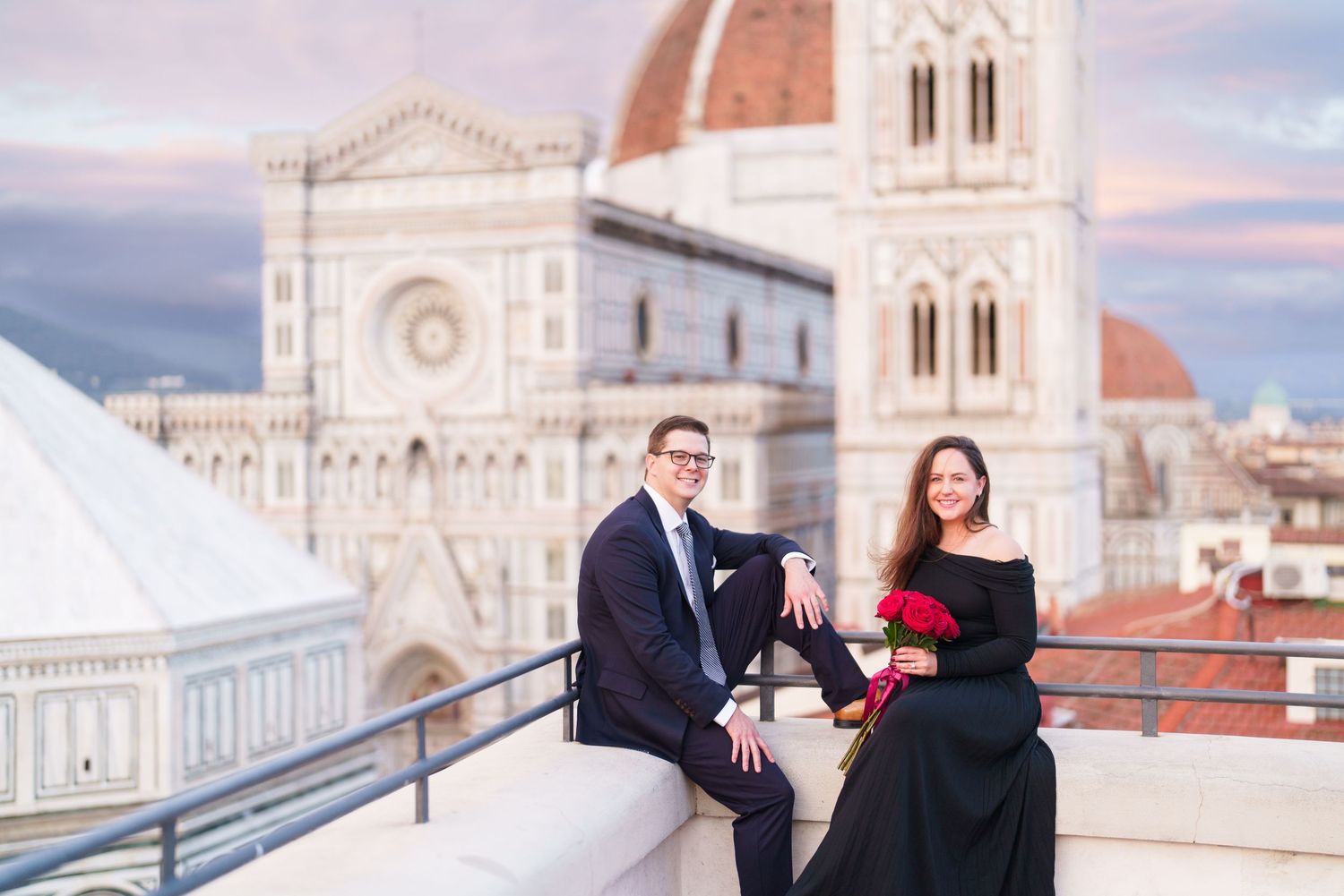 Couple in formal attire sitting on terrace with Florence Cathedral and Duomo in background during sunset.