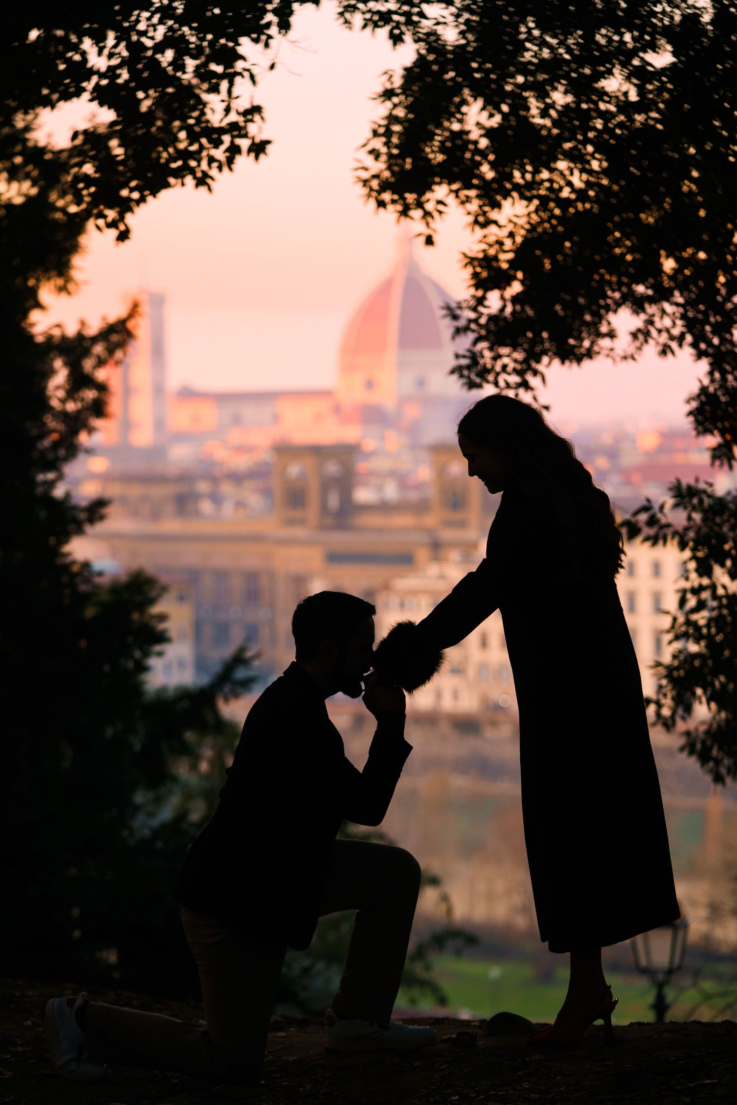 Silhouettes of two people sharing a drink at sunset with trees framing the romantic scene against a city backdrop.