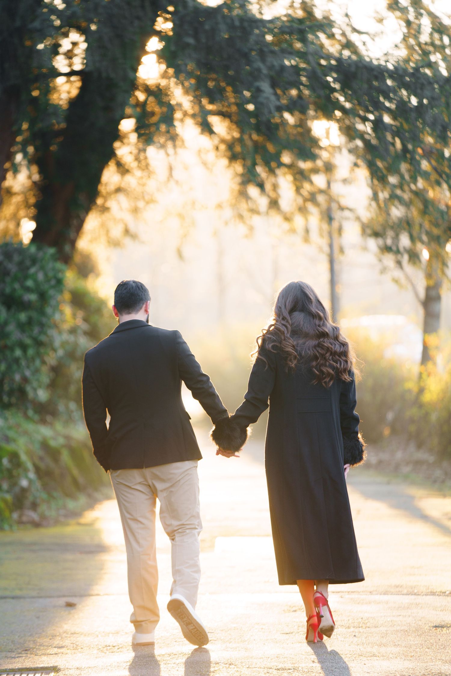 A couple walks hand in hand down a sunlit path through trees during golden hour creating a romantic silhouette.