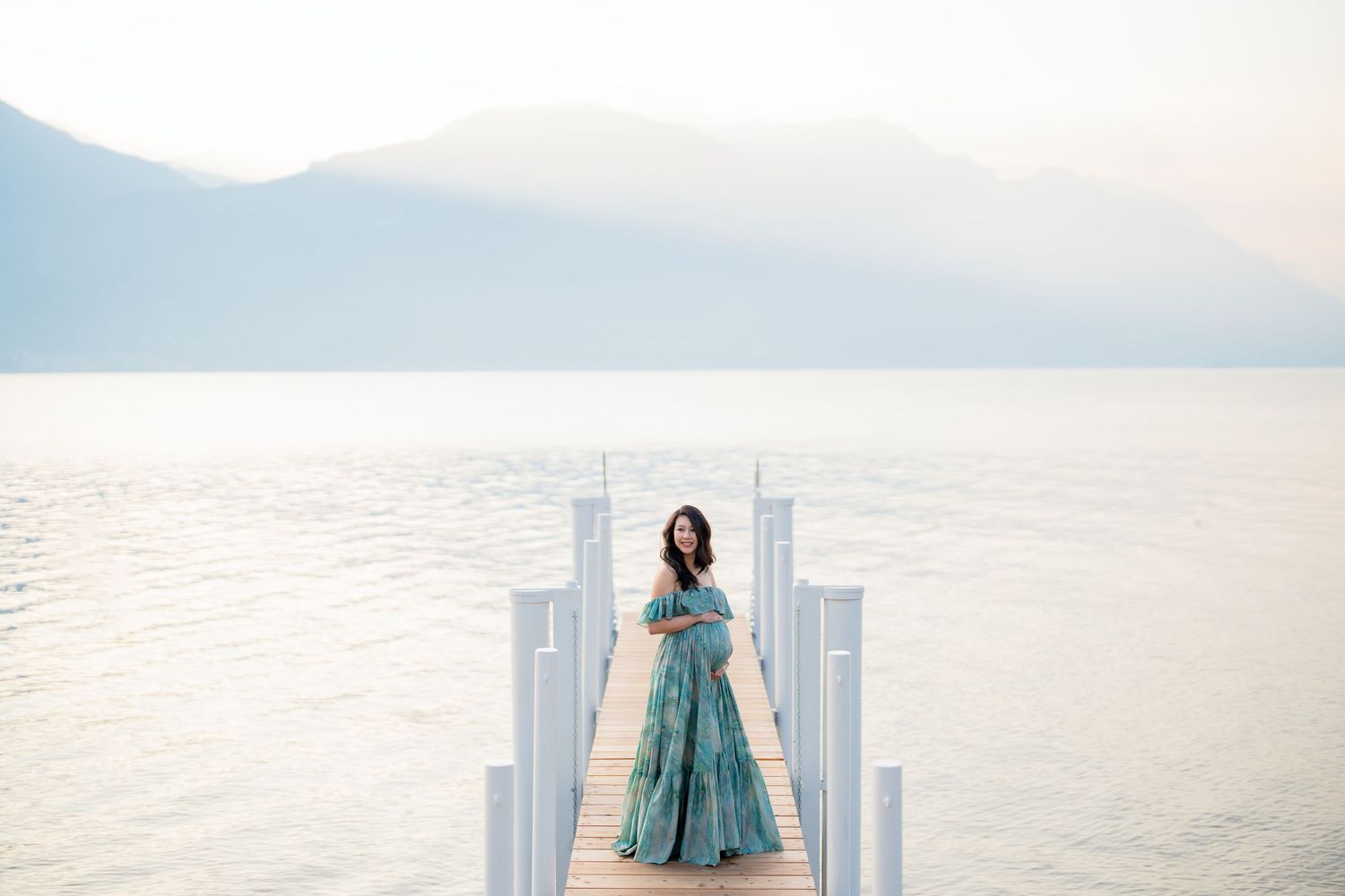 Woman in a flowing turquoise dress poses on a white beach with ocean and mountains in the background.