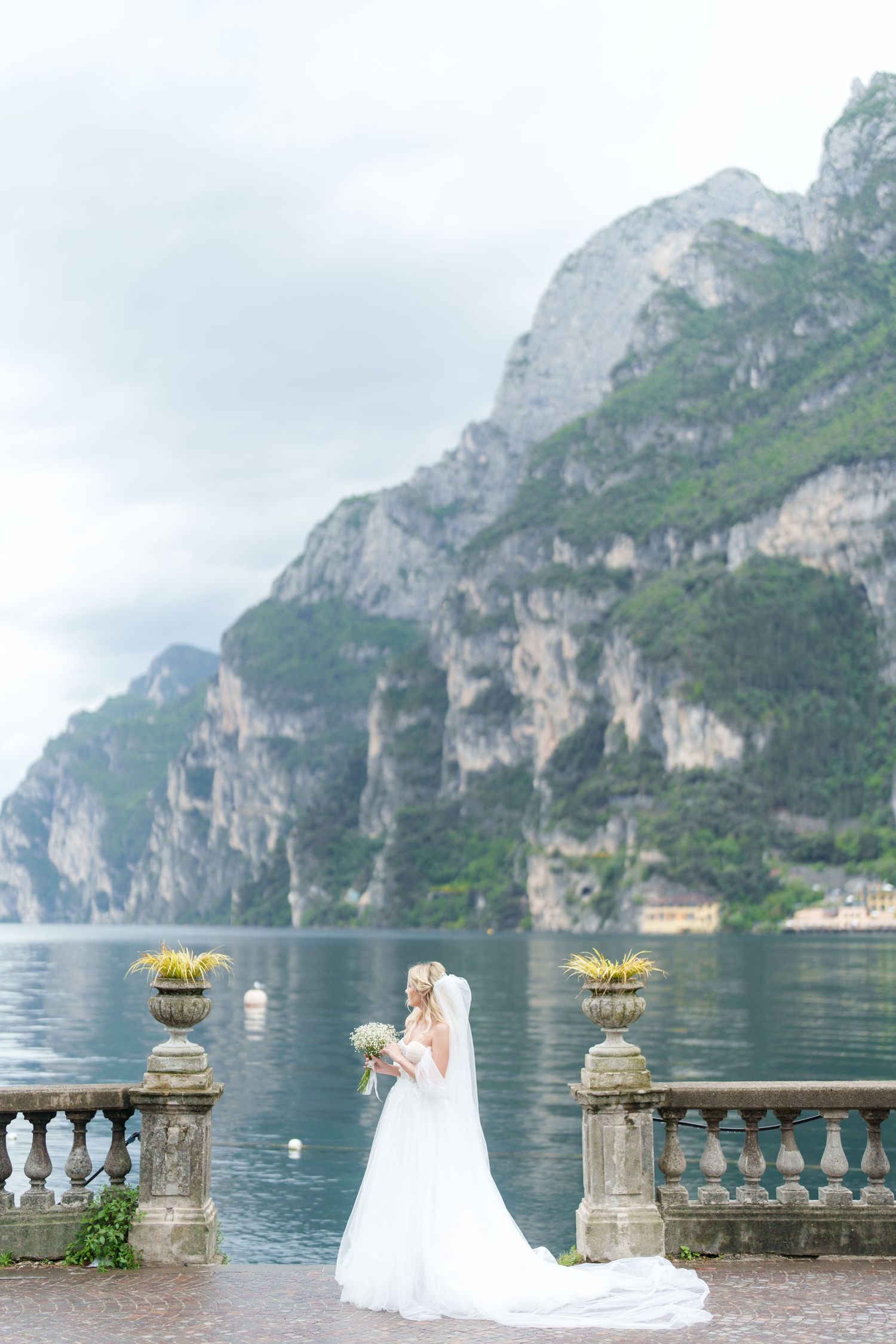 Scenic wedding photo at Lake Garda in Italy with dramatic mountain cliffs and ornate stone balustrade overlooking calm waters.