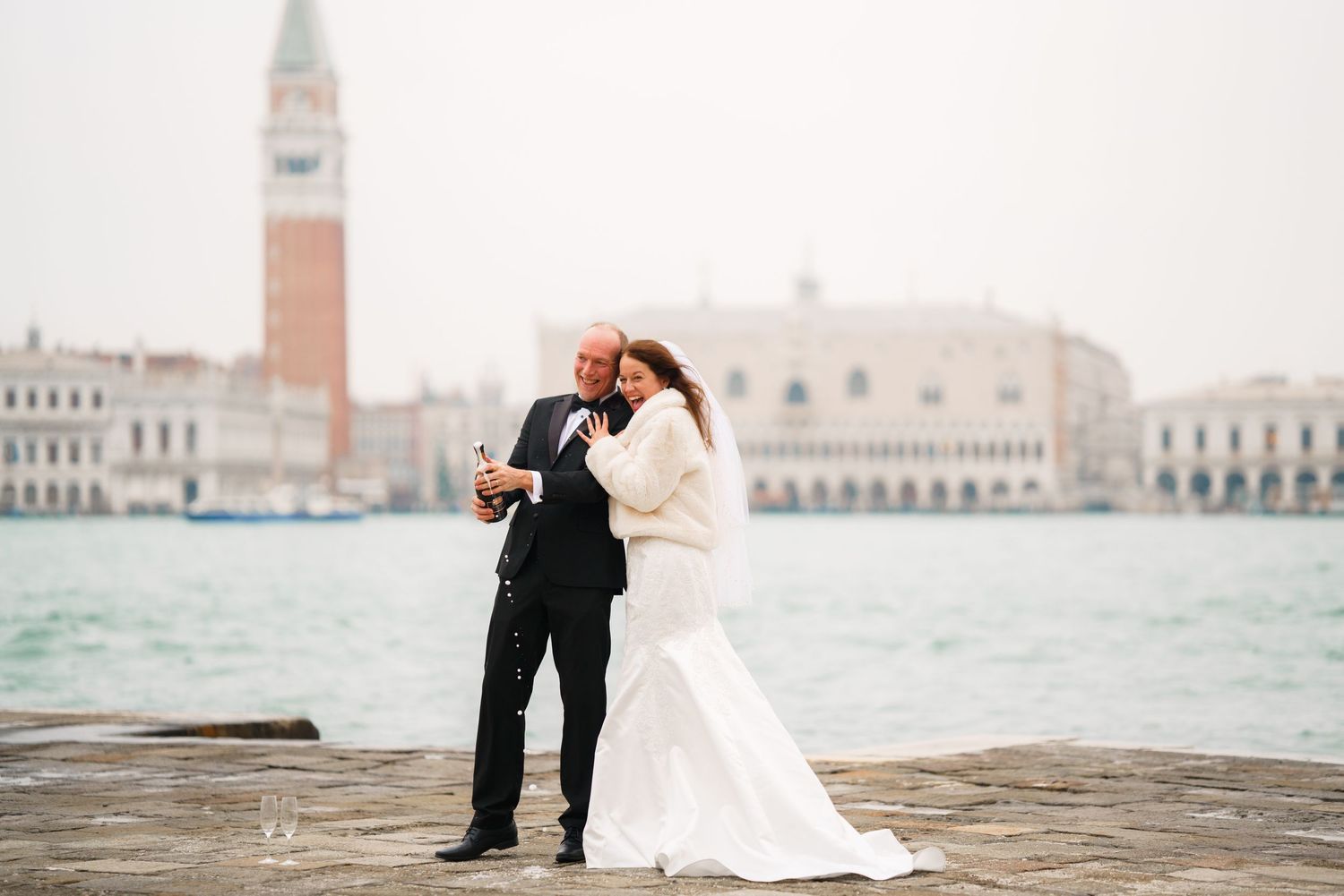 A couple in wedding attire embraces along the waterfront of Venice with historic buildings in the background.
