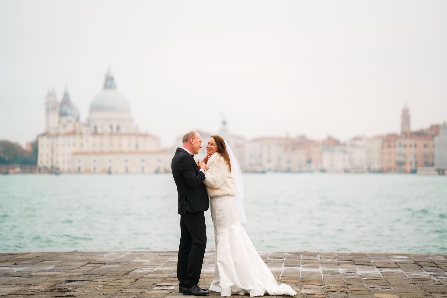 A romantic wedding couple embraces on the waterfront in Venice with the Santa Maria della Salute church in the background.