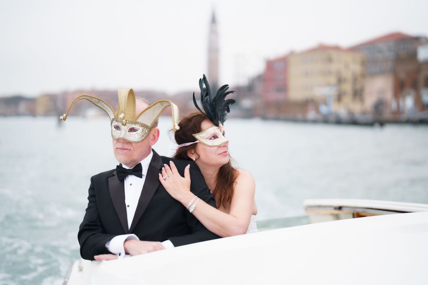 Two people wearing elegant masquerade masks and formal attire pose together on a boat in Venice.