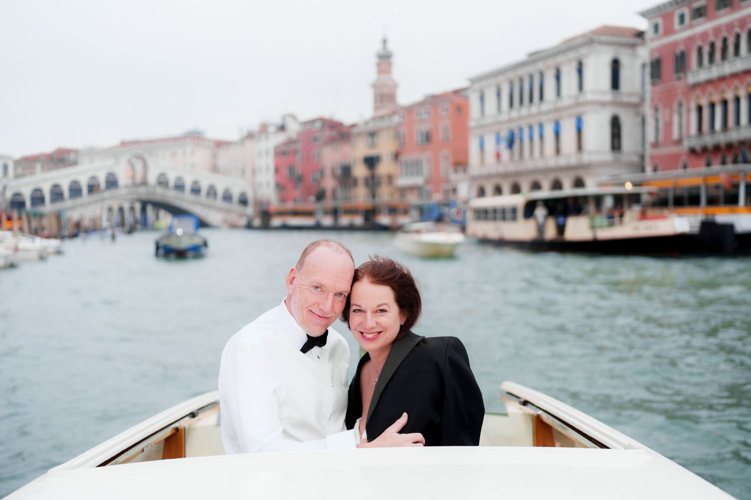 A romantic couple rides in a wooden boat along Venice's Grand Canal with historic buildings in the background.