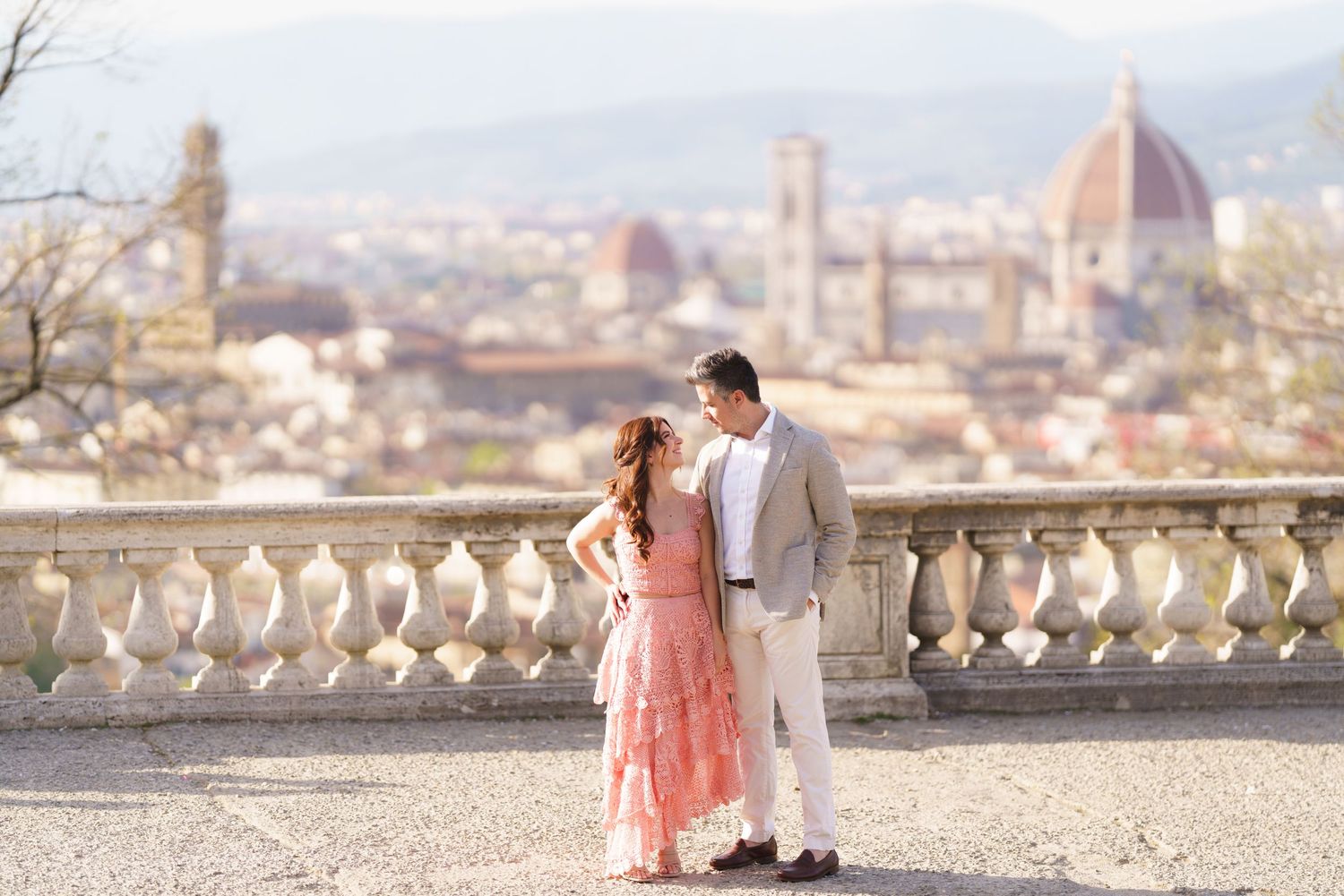 A romantic couple in pink and white outfits stands on a scenic overlook with Florence's iconic Duomo cathedral in the distance.