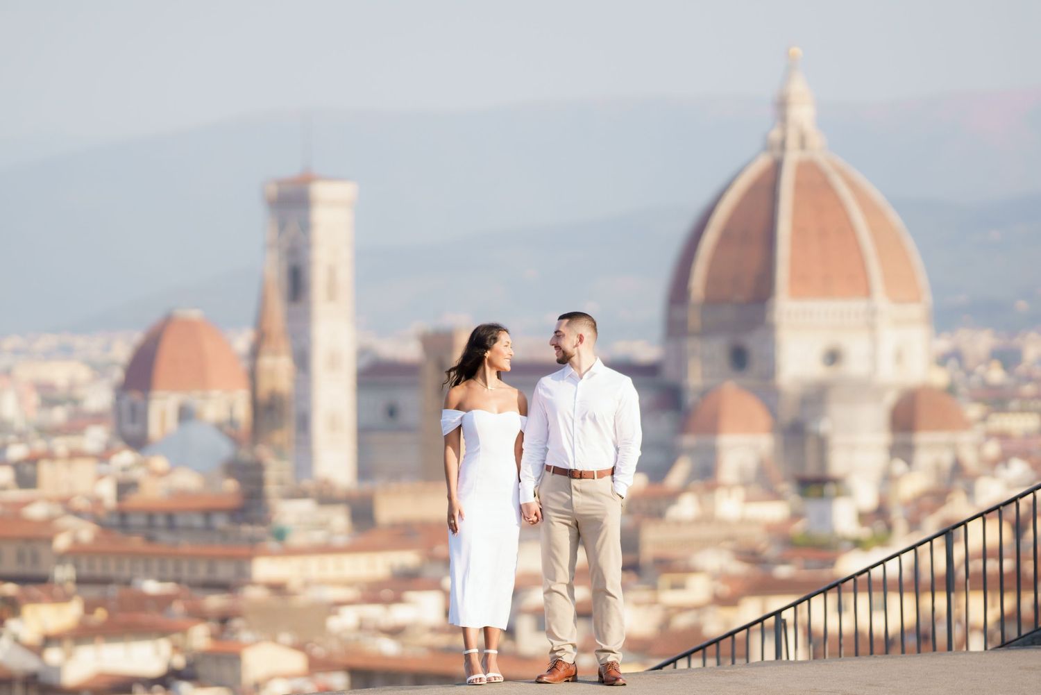 Couple in white outfits walking along Piazzale Michelangelo overlooking Florence's iconic Duomo cathedral at sunset.