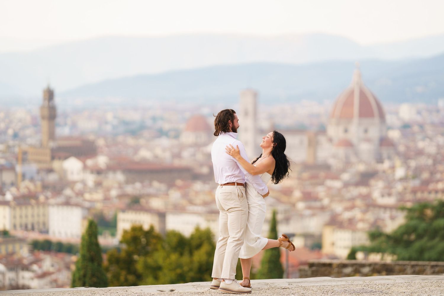 A romantic couple in white outfits embraces while overlooking the scenic cityscape of Florence, Italy with its iconic Duomo.