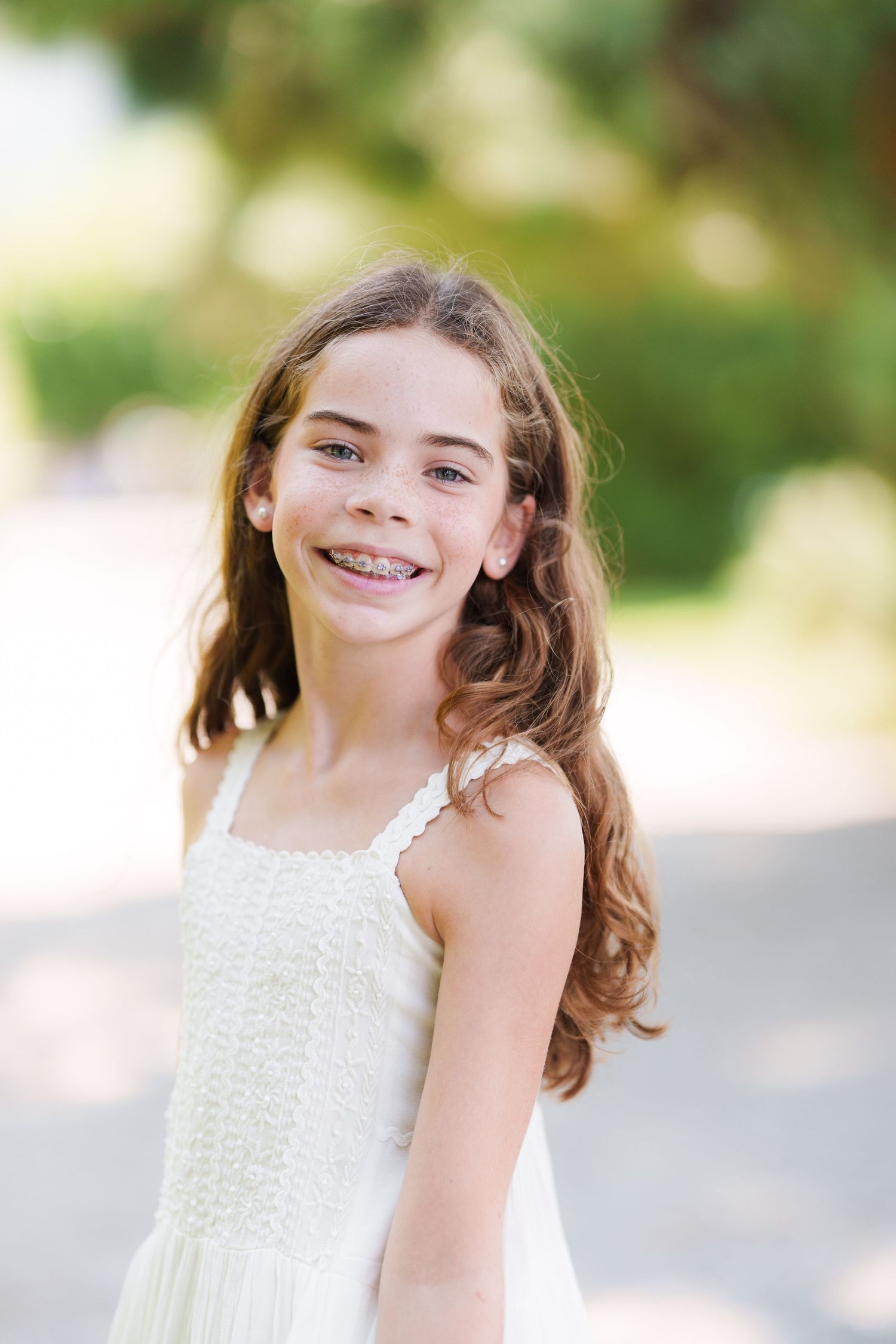 Smiling young girl with braces and long brown hair wearing a white sundress outdoors in natural light.