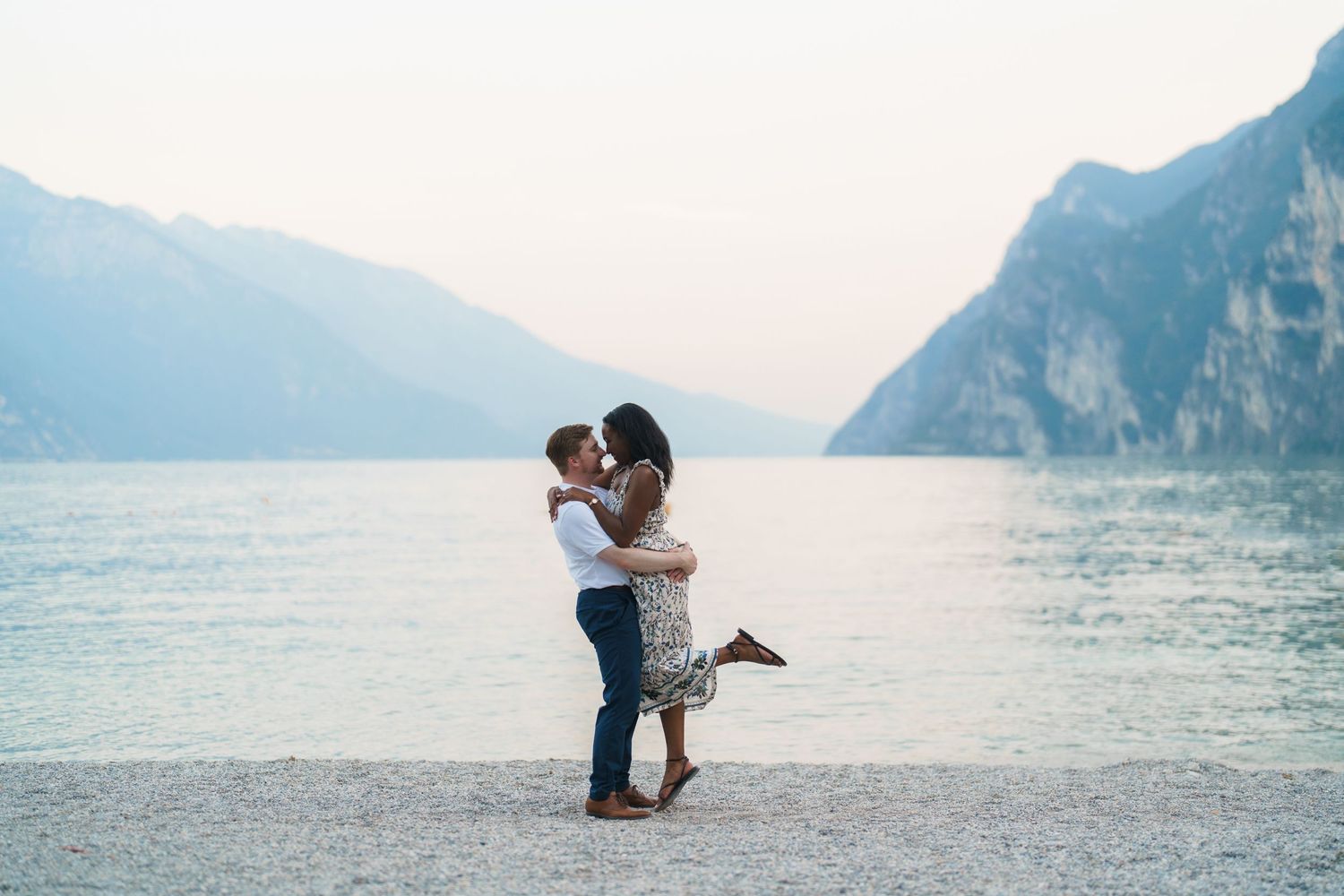 Couple sharing a kiss on Lake Garda pebble beach with towering mountains behind.