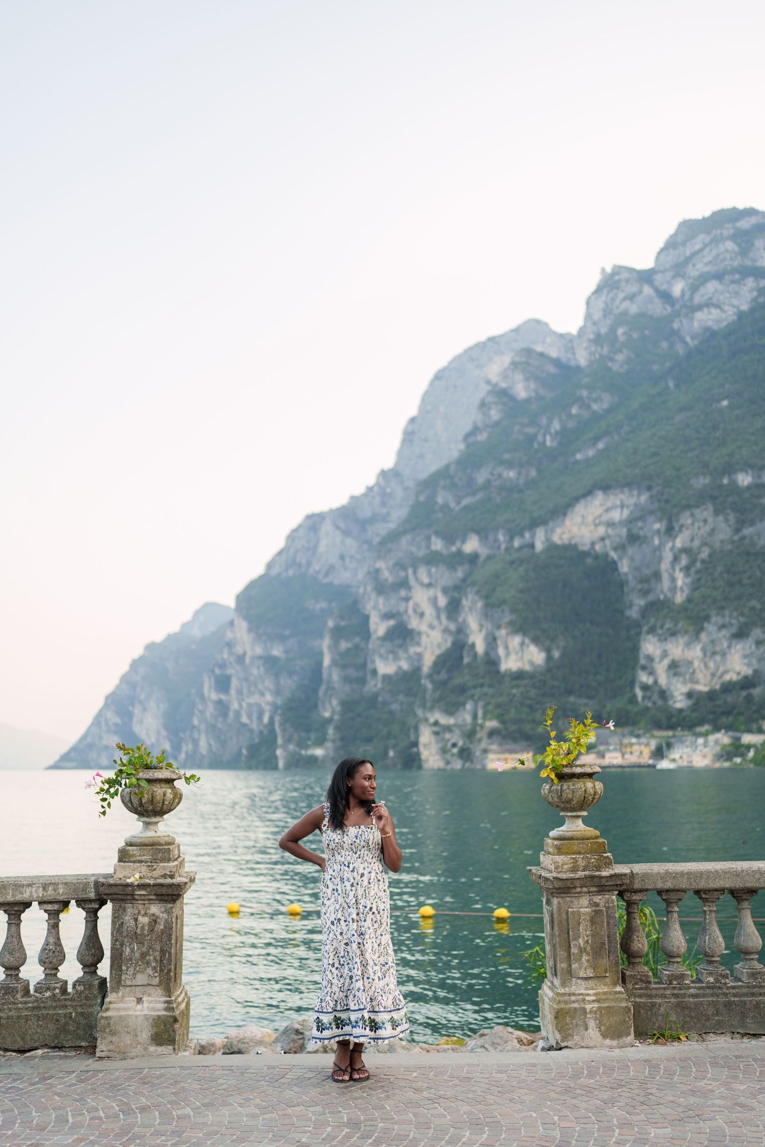 Woman in floral maxi dress standing on scenic Lake Garda terrace with towering rocky mountains backdrop.