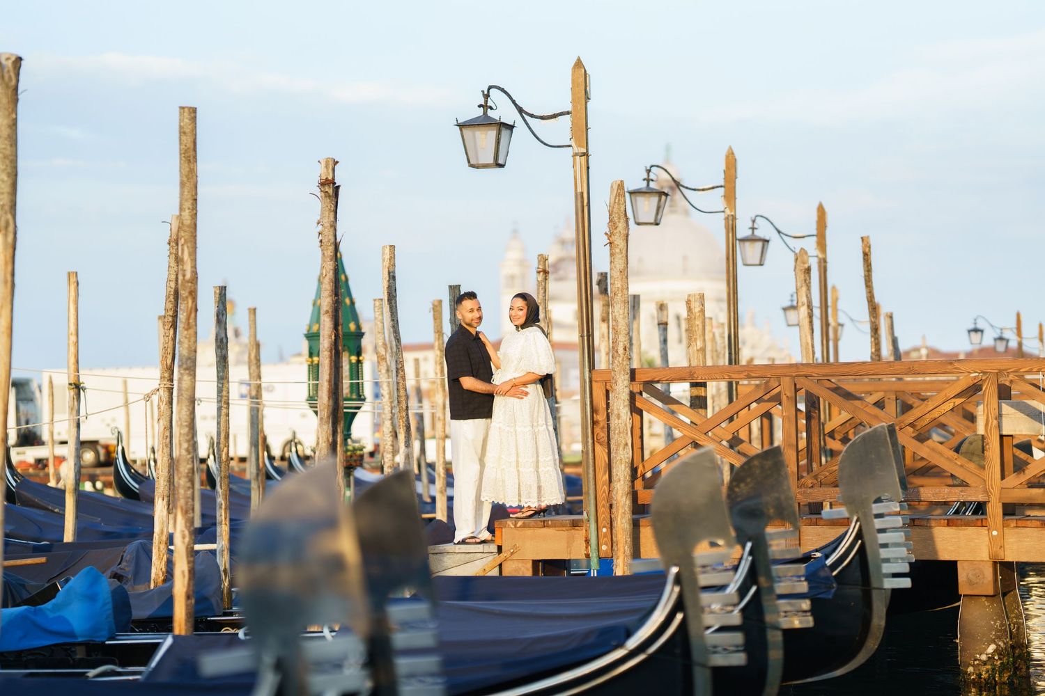 Couple posing romantically among gondolas and wooden poles at Venice waterfront during golden hour.