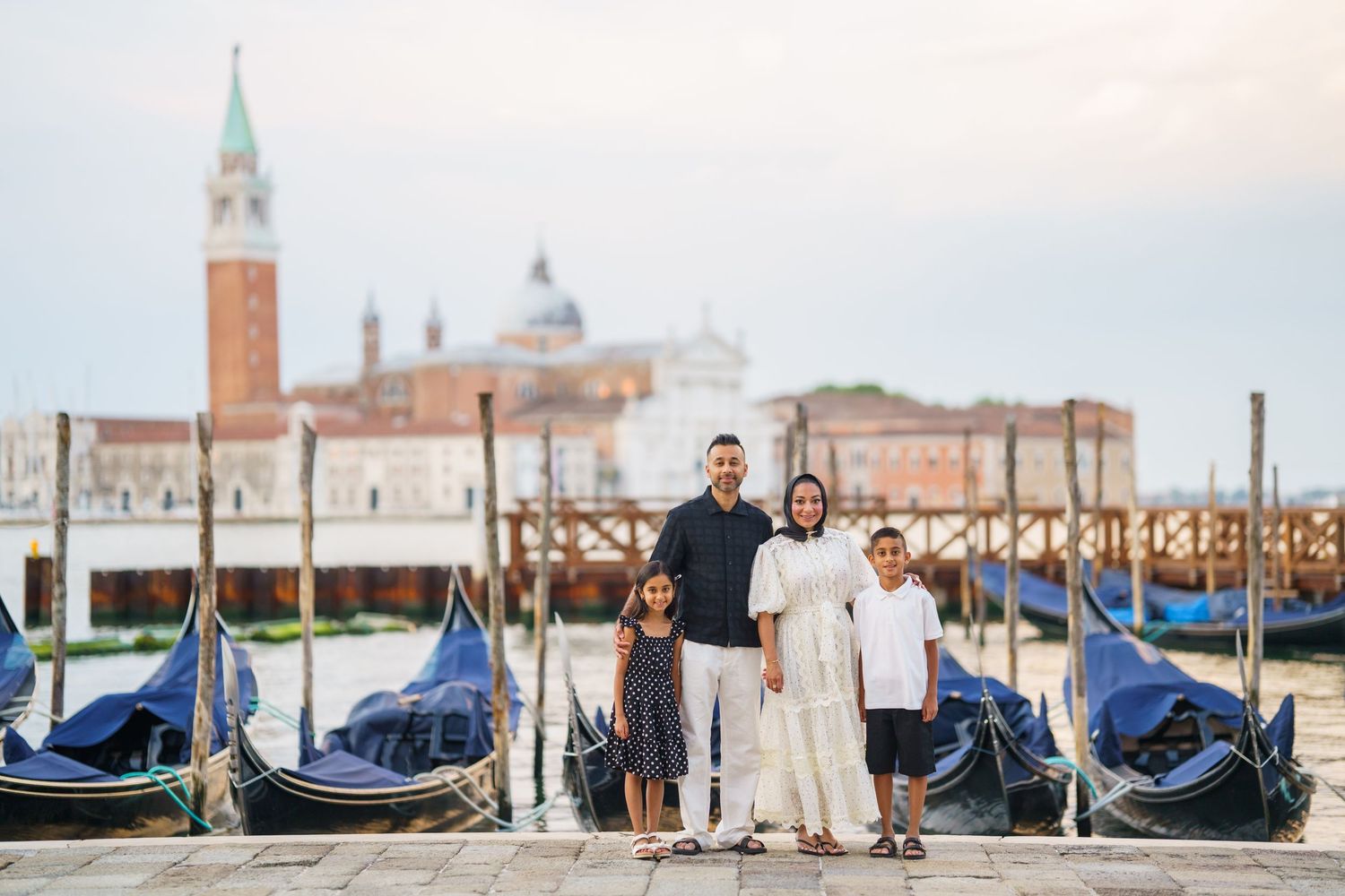 Family of four posing by gondolas on Venice waterfront with San Giorgio Maggiore church in background.