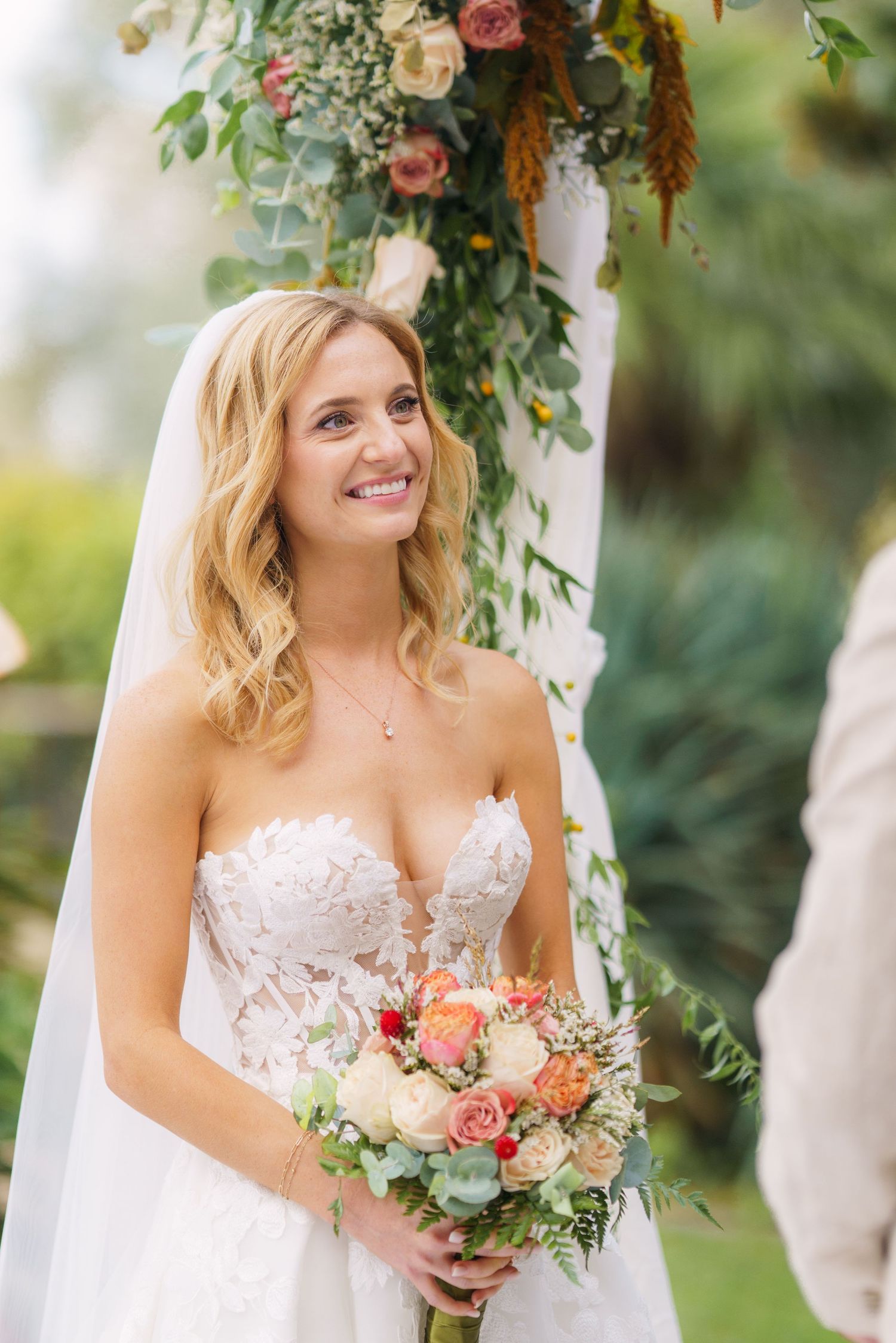 A joyful bride holding a peach and pink bouquet under a floral-decorated wedding arch.