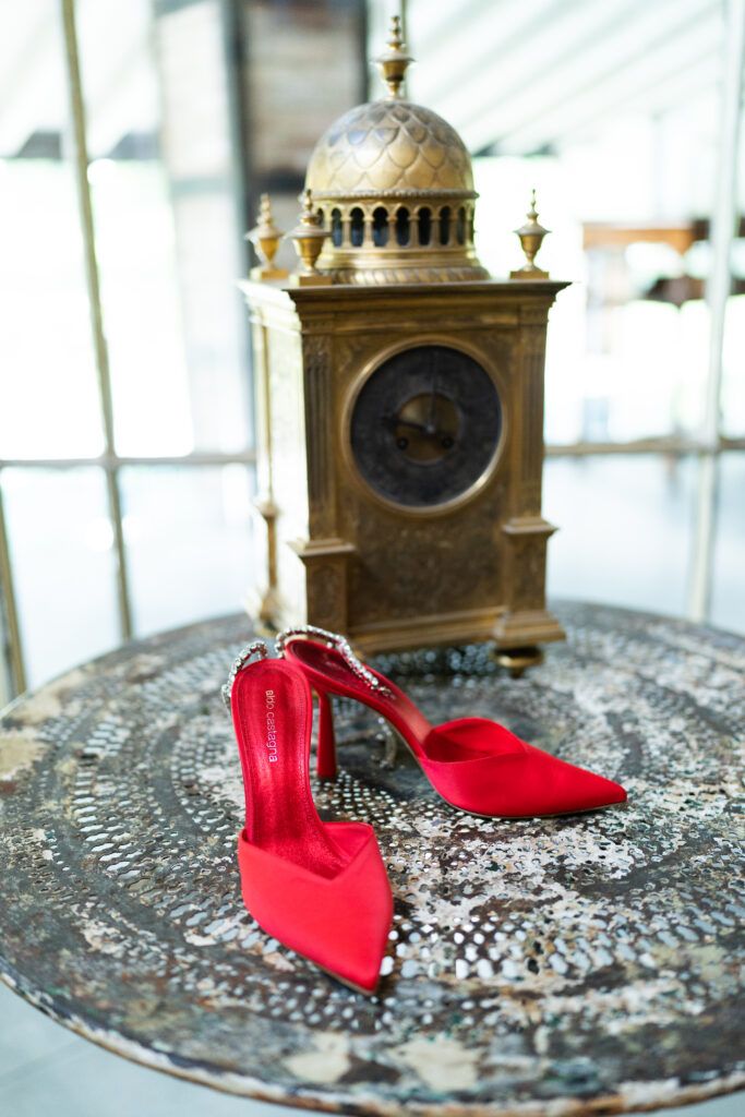 Red high heel shoes displayed next to vintage brass clock on metallic patterned surface.