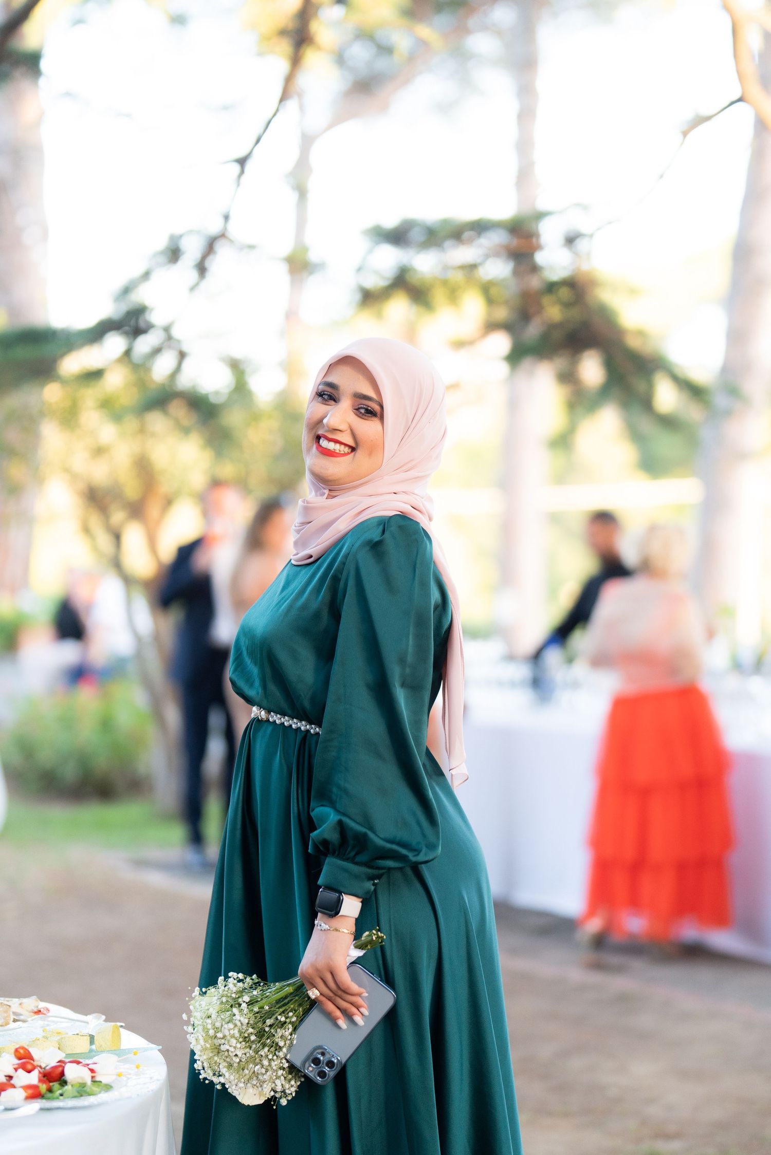 Wedding guest in an elegant emerald green dress and light pink hijab poses with flowers outdoors.