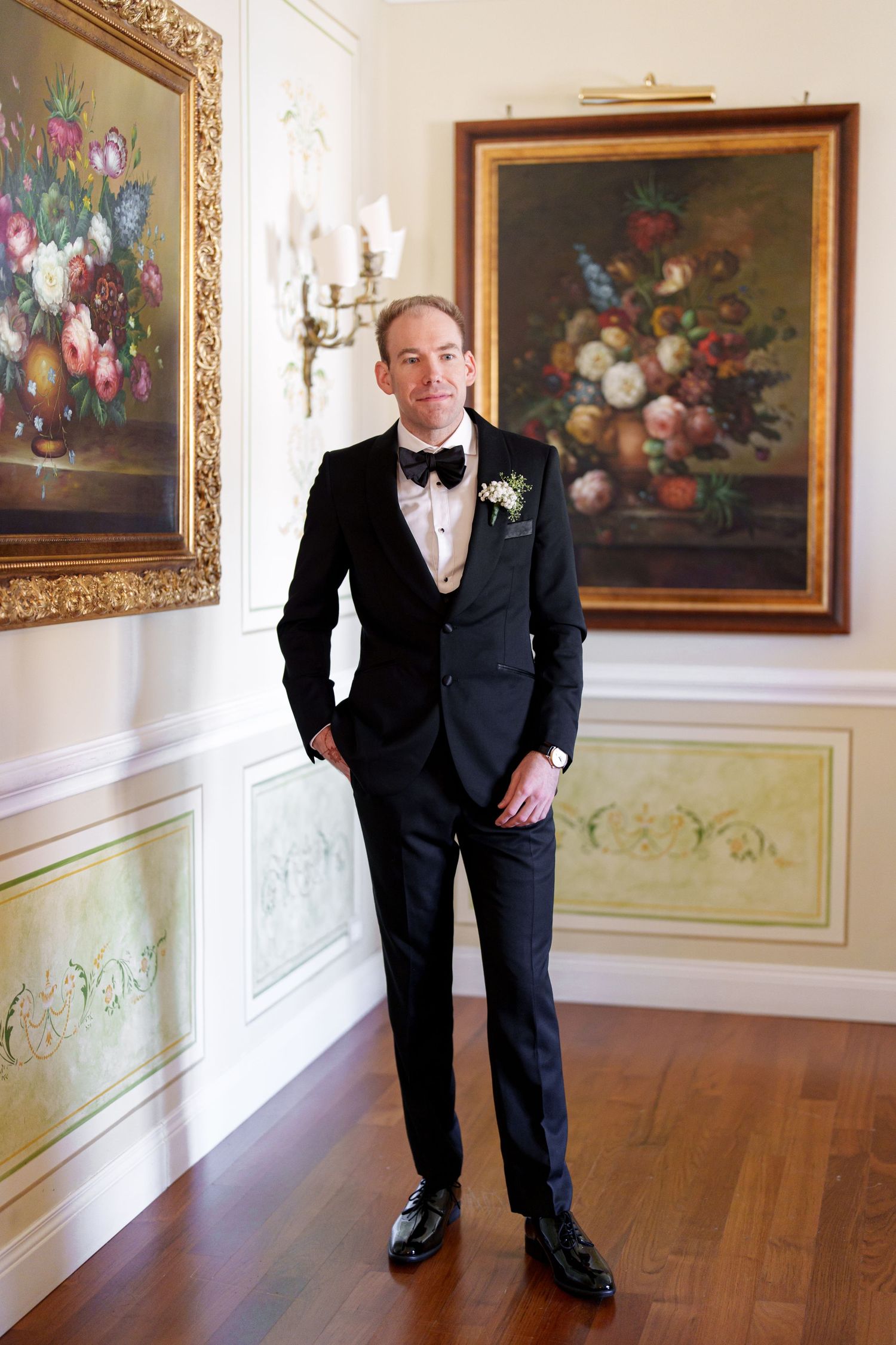 A person in a black tuxedo poses confidently in an elegant hallway with hardwood floors.