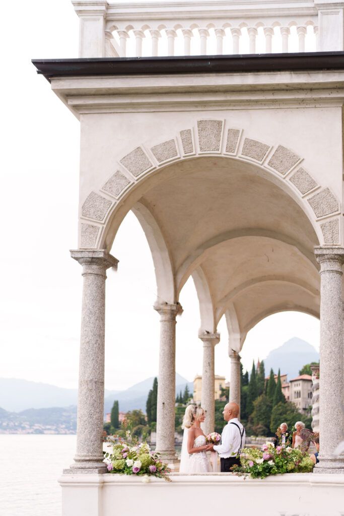 Elegant wedding ceremony under white stone arches overlooking a lake and mountains in Italy.