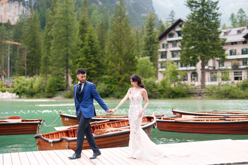 Newlyweds pose by wooden boats on mountain lake dock with resort buildings visible.
