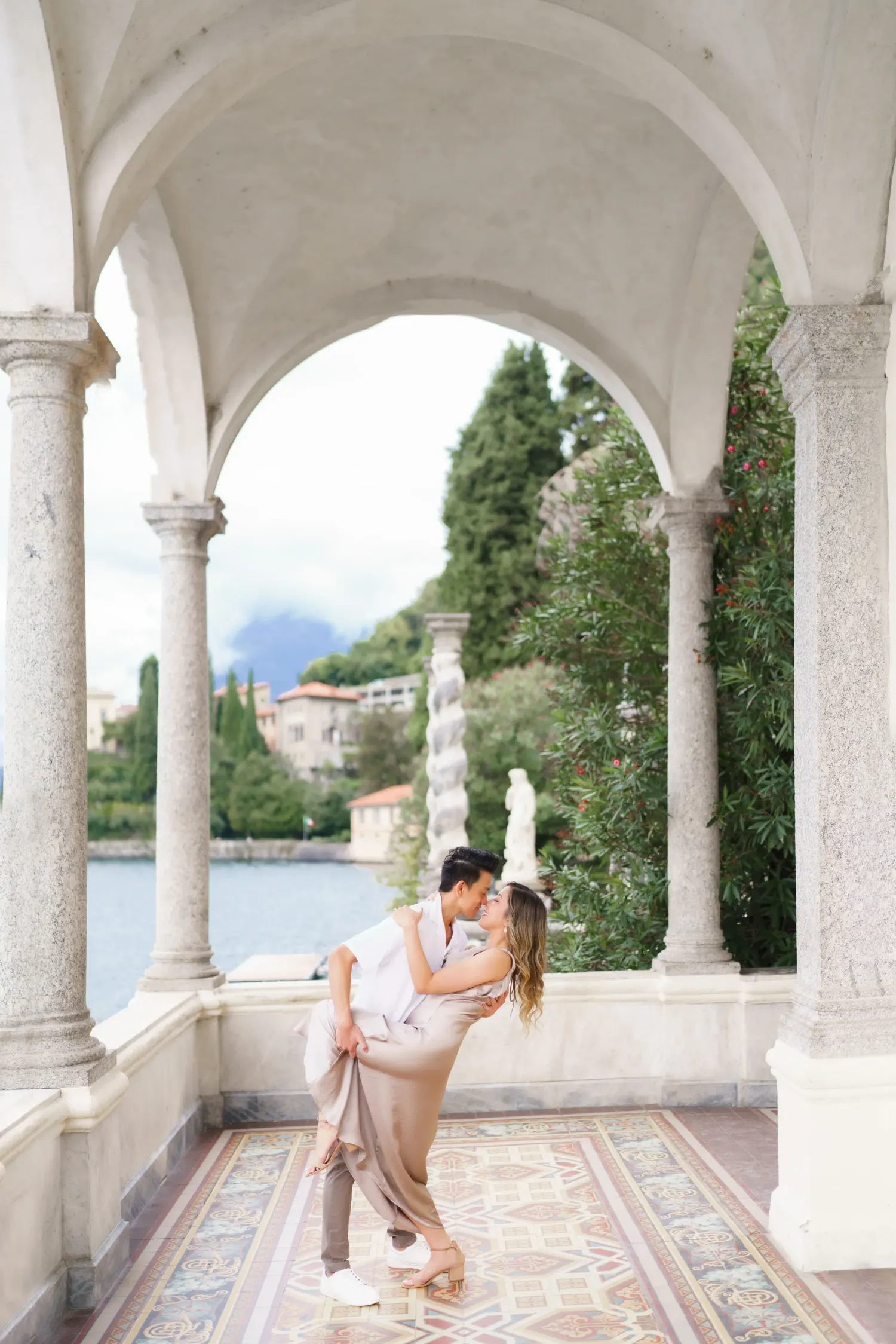 A romantic couple stands under white arched columns overlooking Lake Como in Italy on a sunny summer day.