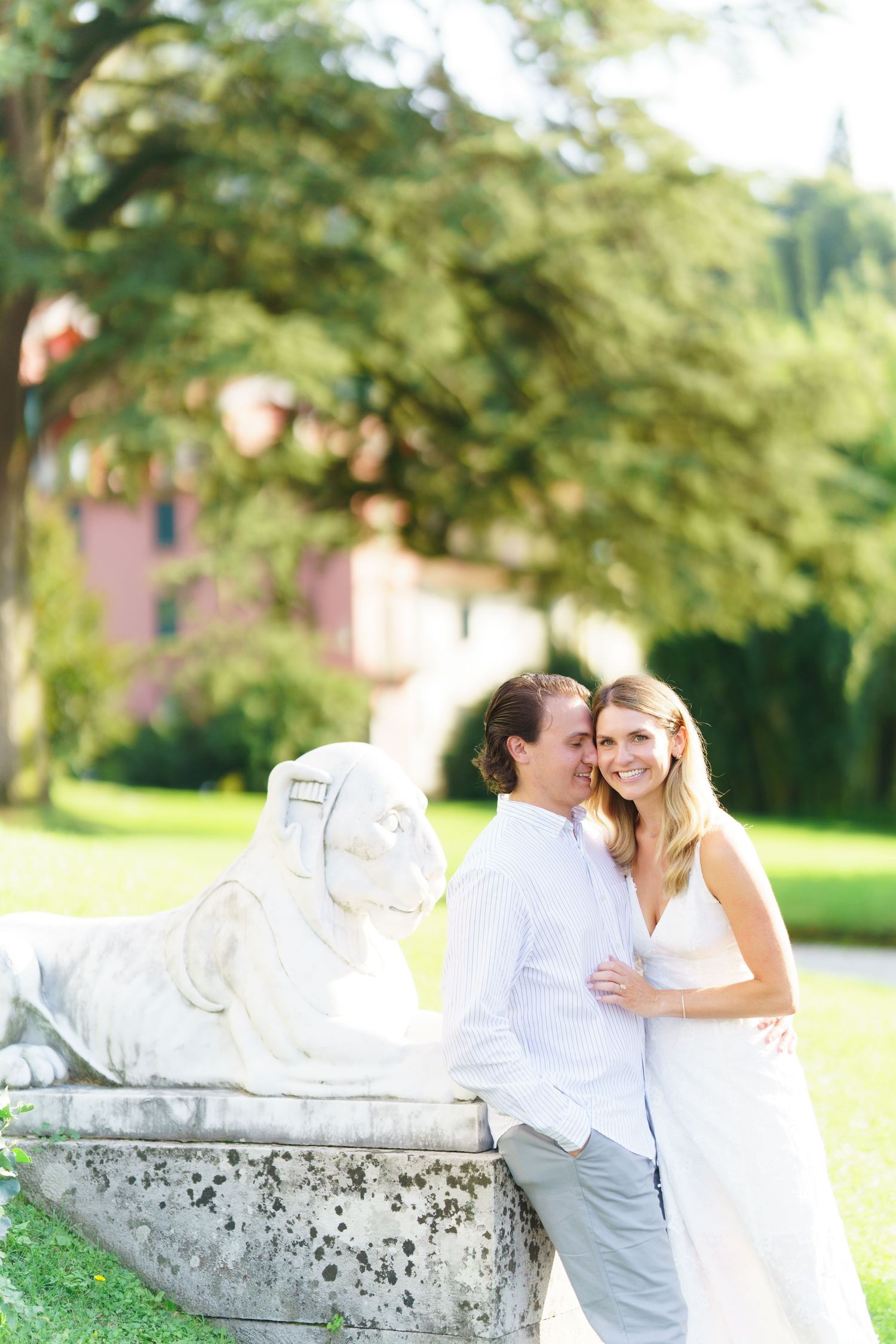 A couple in white clothing sits together on a stone bench in a lush garden setting during a romantic photoshoot.