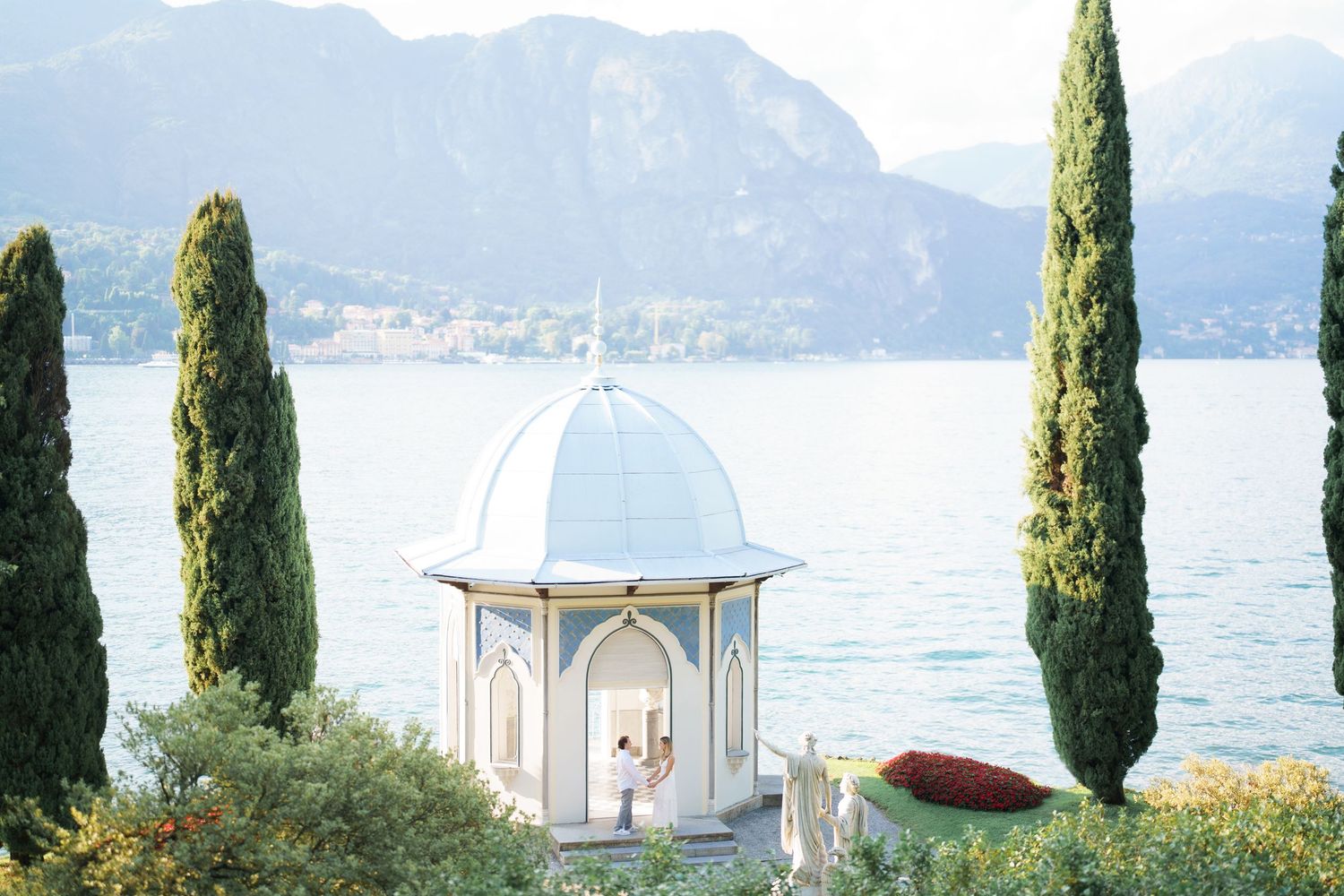 Scenic Lake Garda view with cypress trees and white gazebo along the waterfront promenade in Italy.