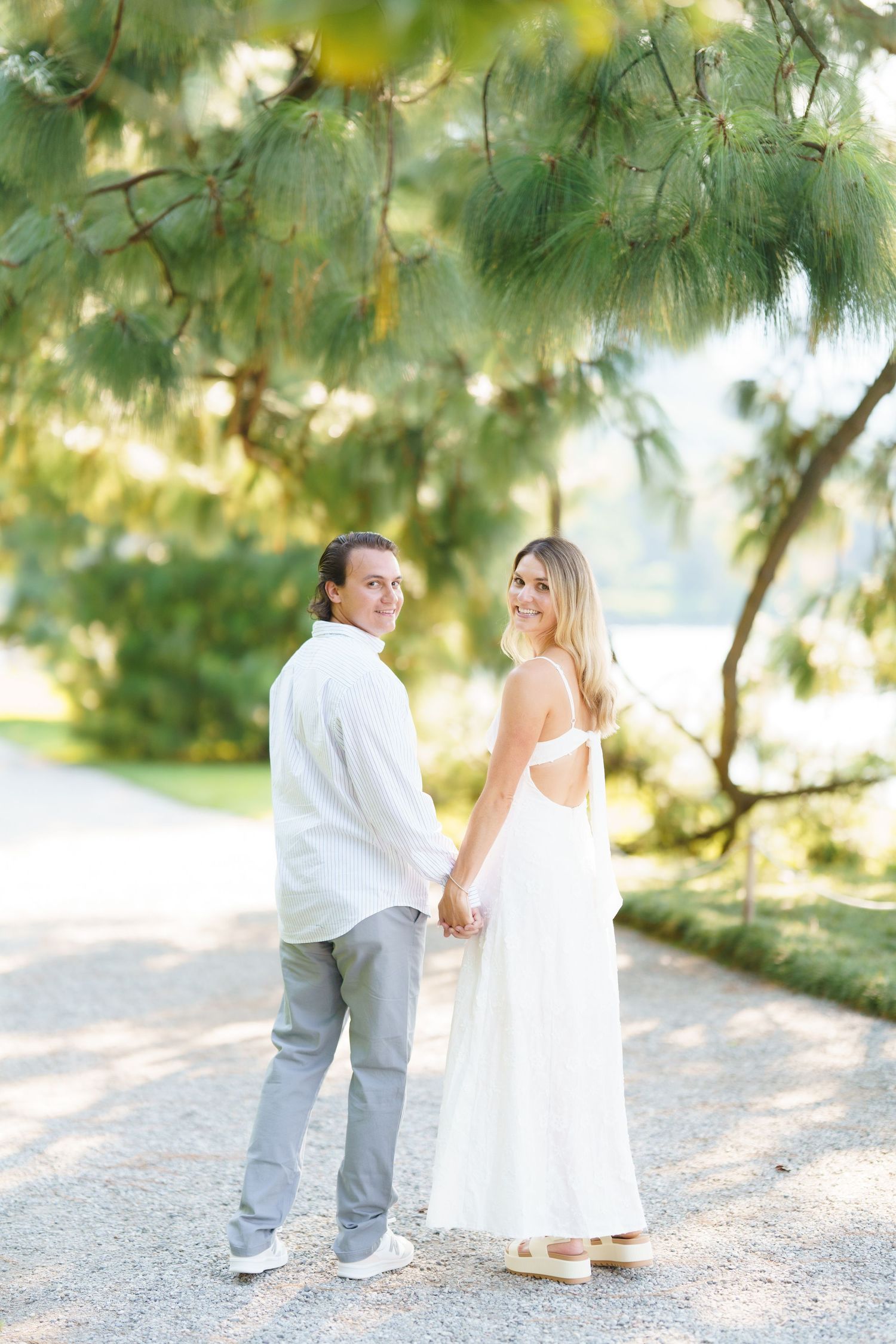 A couple holds hands on a sunlit path under overhanging trees in a romantic outdoor setting.