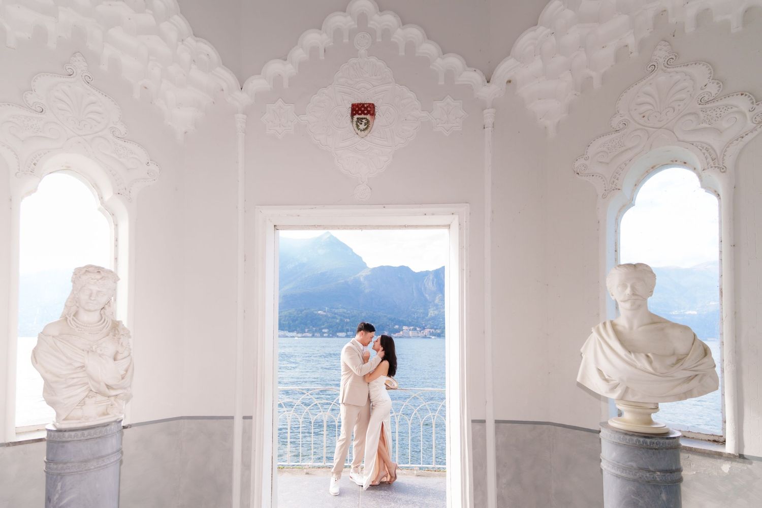 A romantic couple poses in a white Mediterranean-style archway overlooking a scenic coastal view.