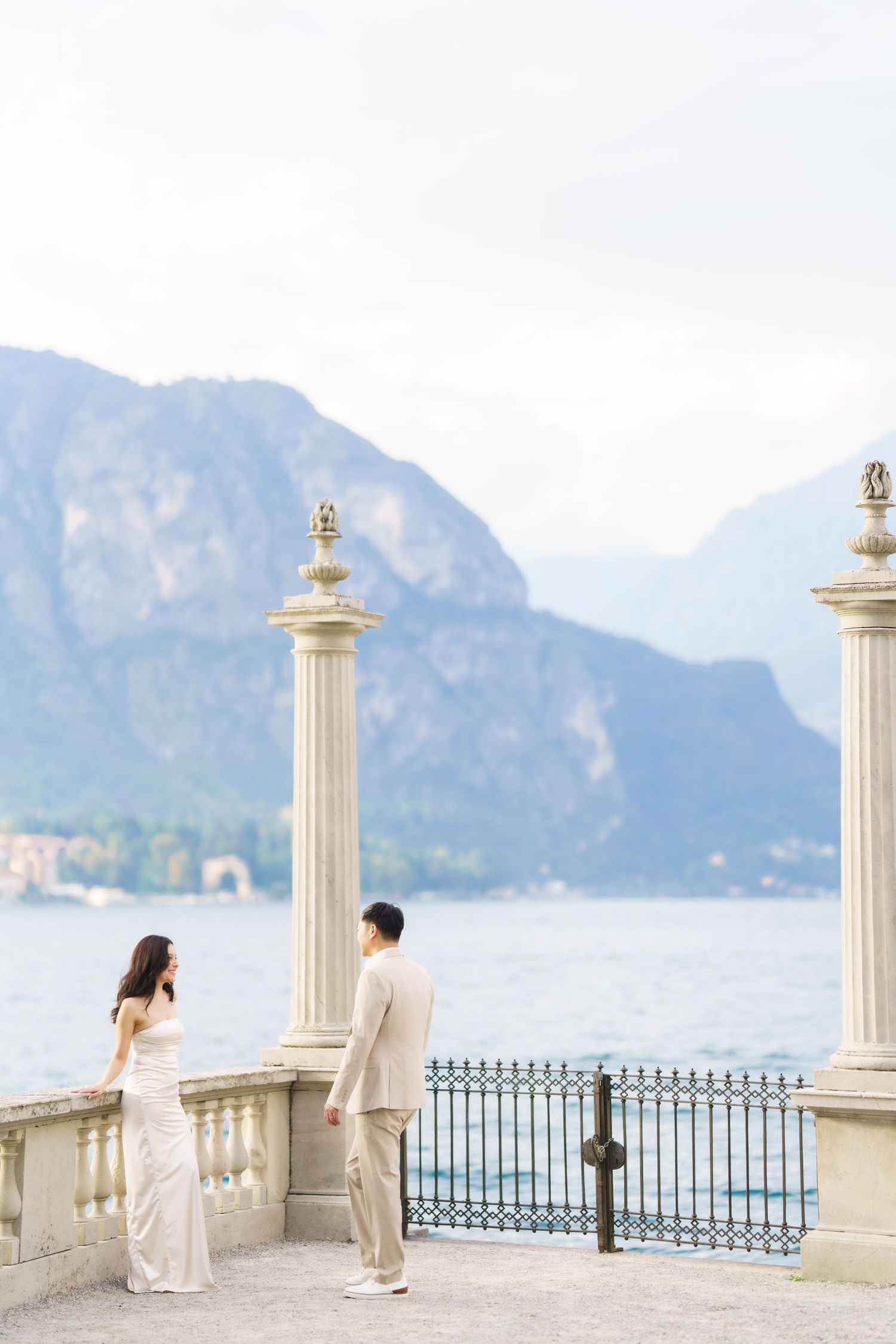 A couple shares a romantic moment on an elegant balcony with white columns overlooking a scenic mountain lake view.