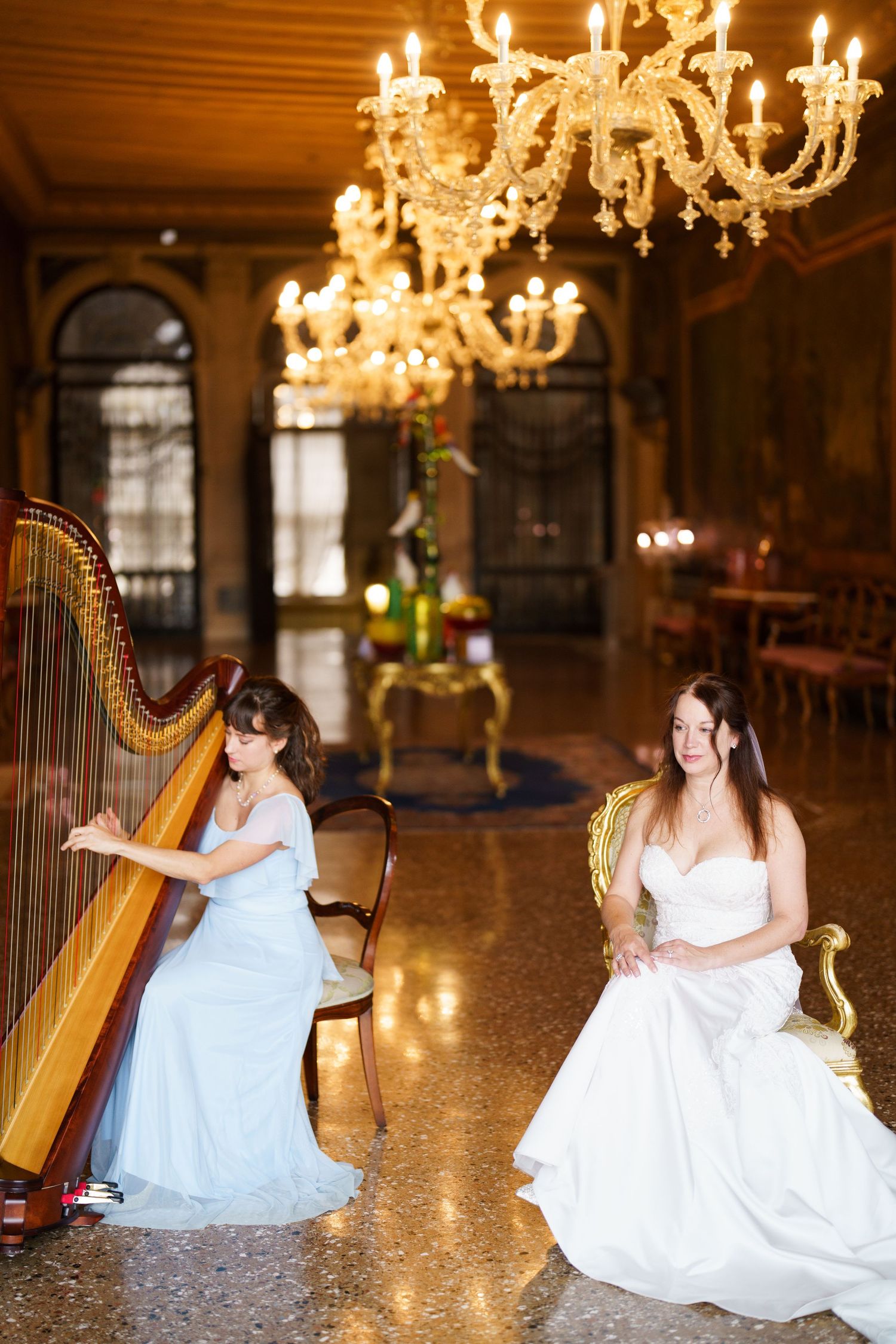 A harpist in an elegant gown performs in an ornate ballroom with crystal chandeliers and polished marble floors.