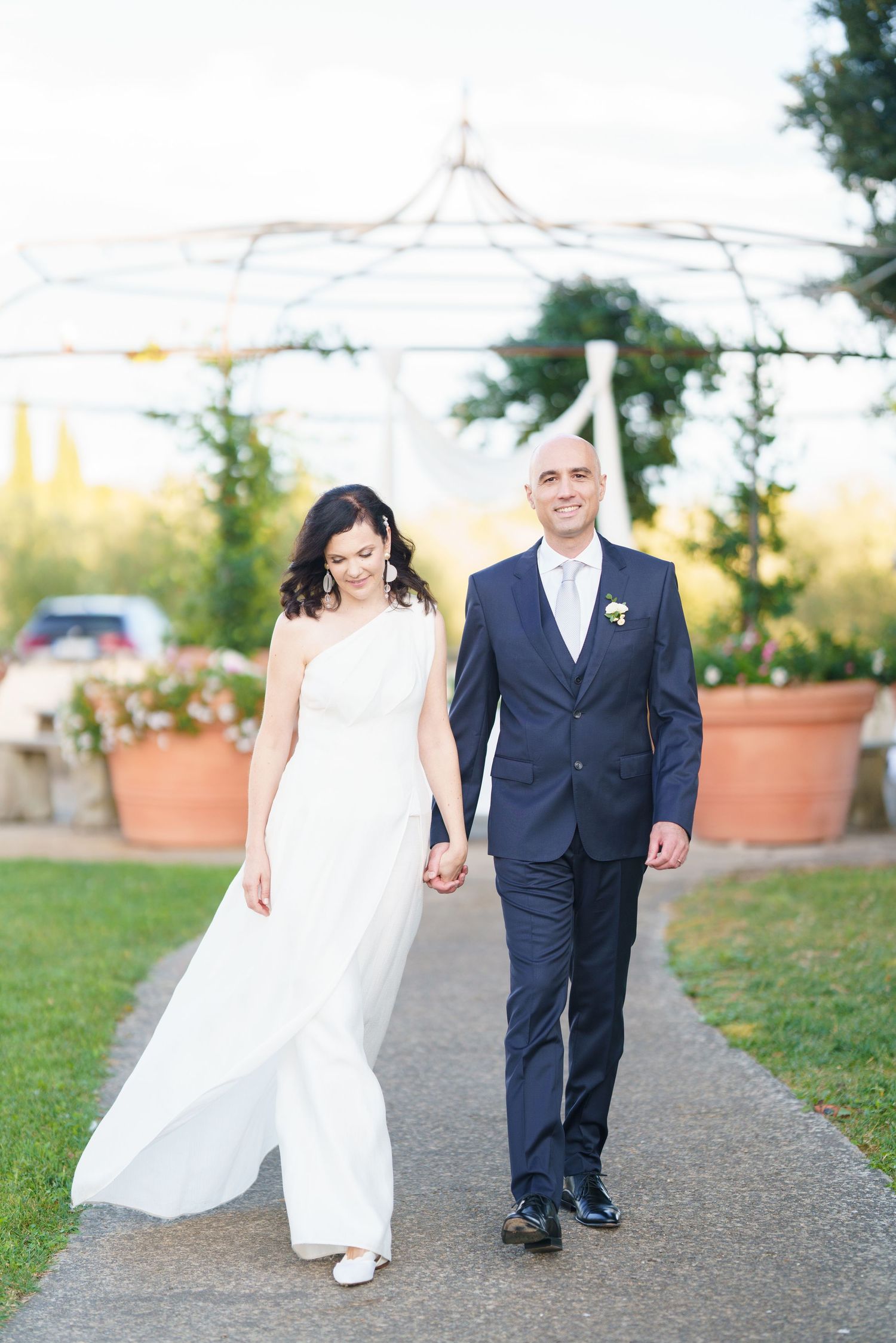 Romantic wedding couple walking hand in hand on outdoor path with potted plants.