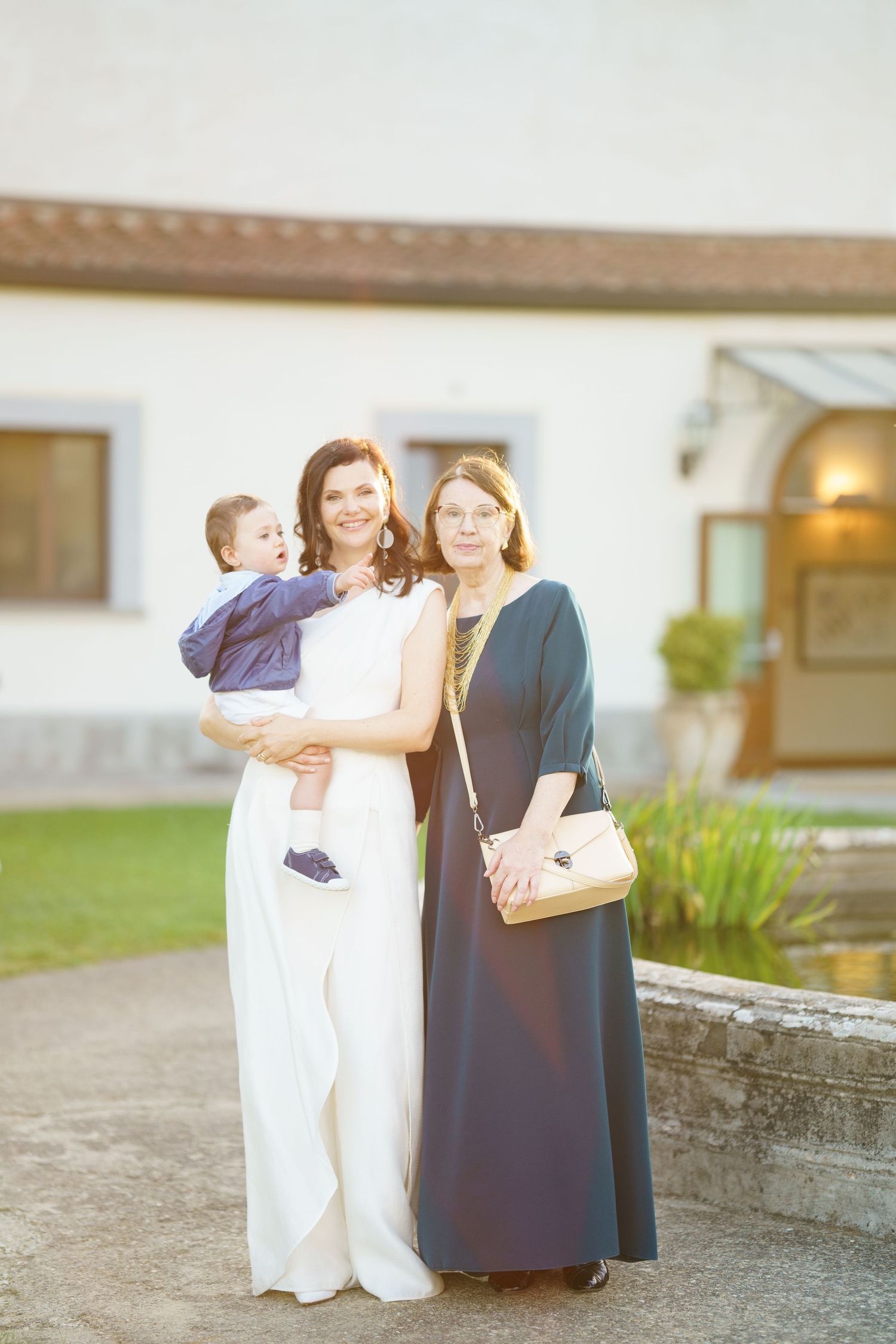 Three people in formal attire pose together outside a white building in evening light.