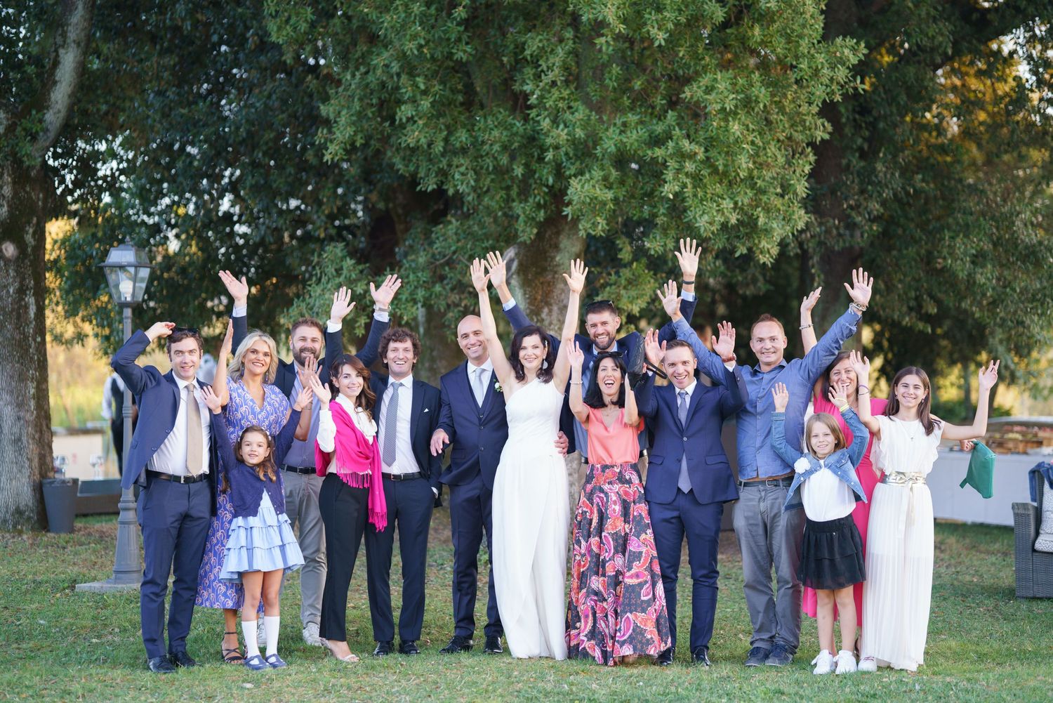 Joyful wedding party raises hands in celebration pose during outdoor group photo.
