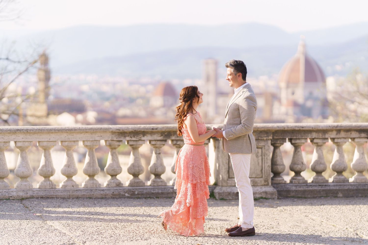 A romantic couple stands on a scenic overlook terrace with Florence's iconic Duomo cathedral visible in the background.