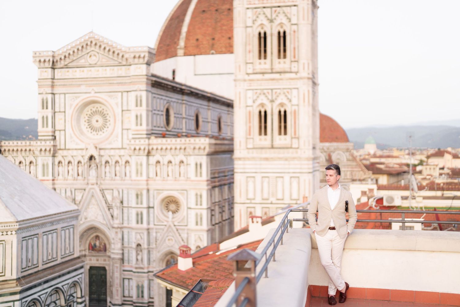 View of Florence Cathedral's ornate facade and bell tower with person standing on rooftop terrace overlooking city.
