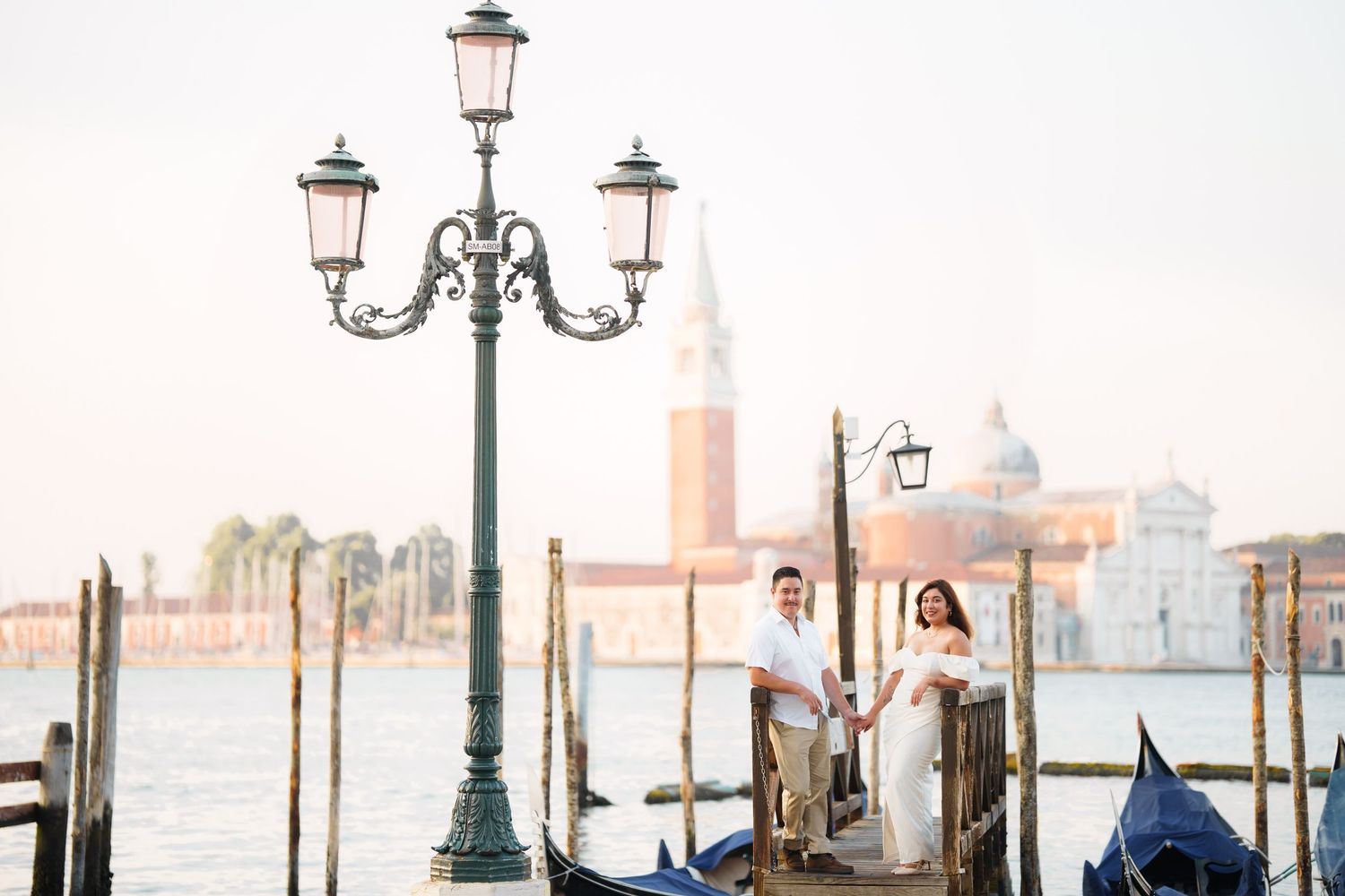 Couple stands beside ornate lamp post on Venice waterfront with gondolas and San Giorgio Maggiore church in background.