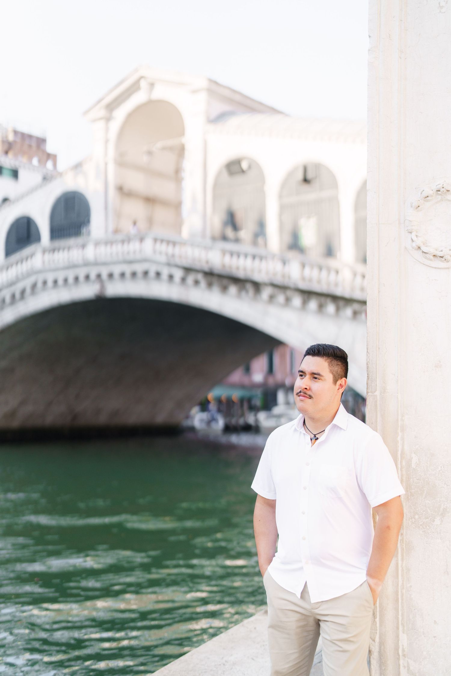 Man in white shirt poses along Venice canal with Rialto Bridge in background during sunny day in Italy.
