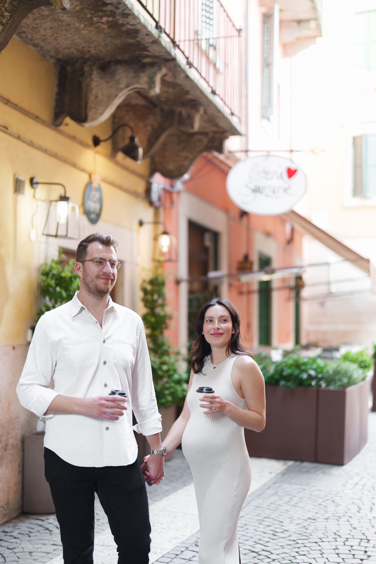 Couple in white outfits walking through European cobblestone street while looking at mobile phones.