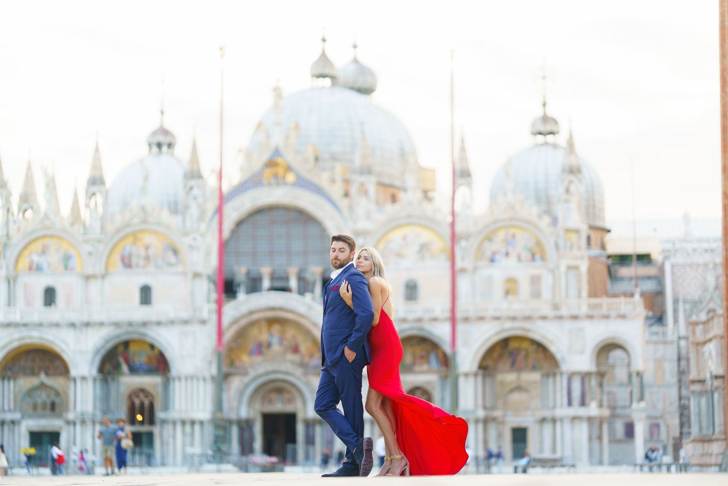 A couple in formal attire stands in front of St. Mark's Basilica in Venice with a flowing red dress catching the wind.
