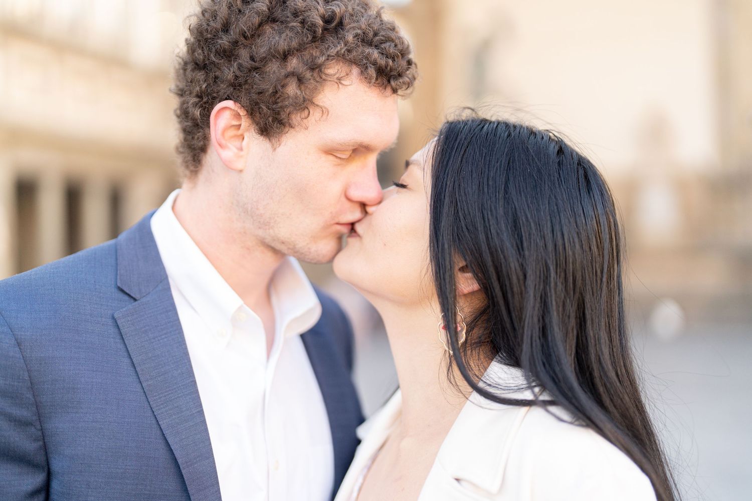 A couple shares a romantic moment in an elegant outdoor setting with blurred architectural details in the background.