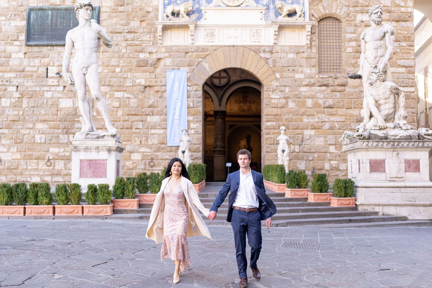 A couple walks hand in hand through a historic Italian plaza with Renaissance statues and stone architecture in Florence.