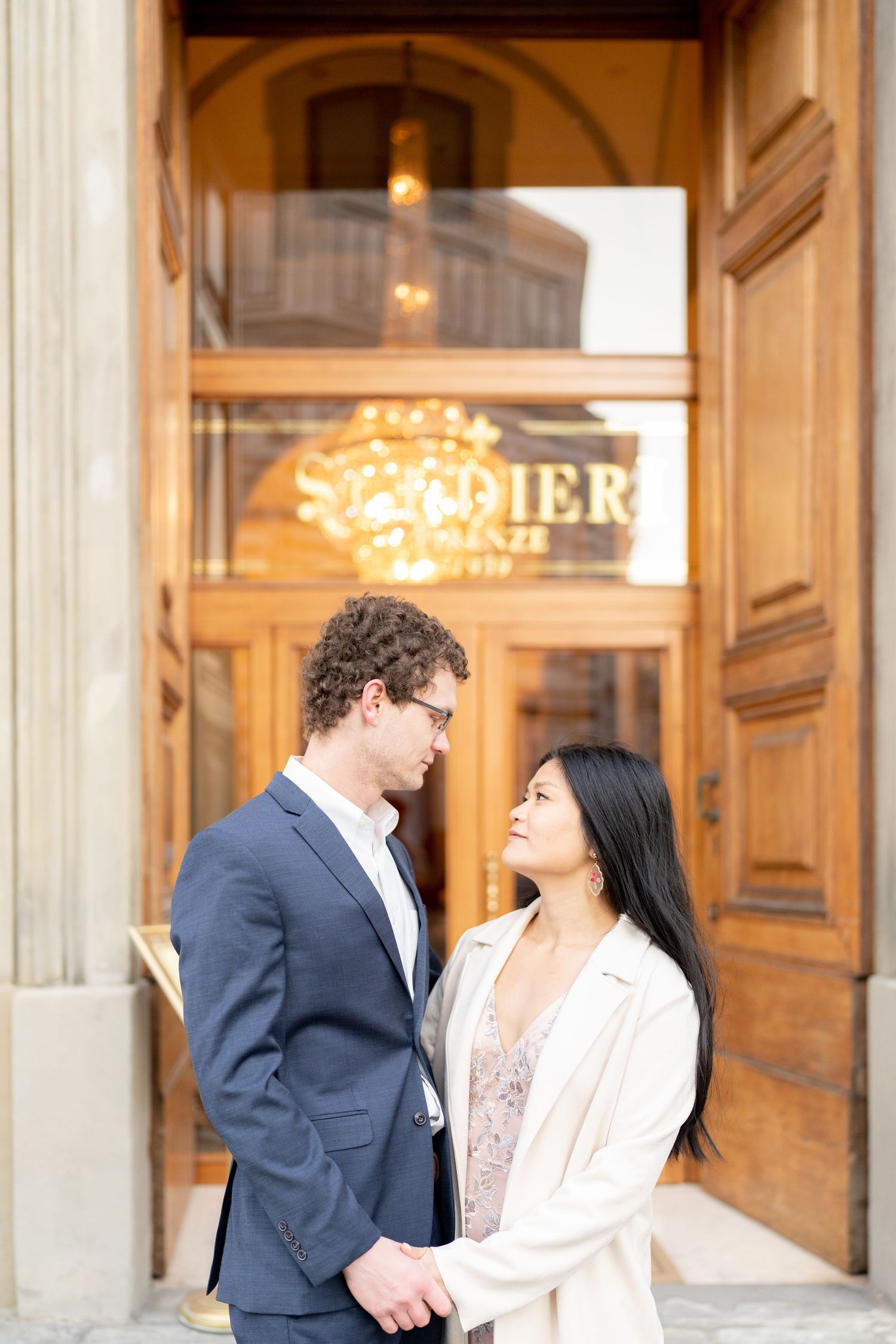 A couple shares an intimate moment in front of an elegant building entrance with ornate wooden doors and warm lighting.