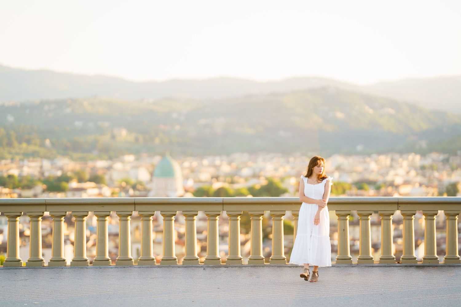A person in white dress walking along a scenic waterfront with bridges and mountains in the background.