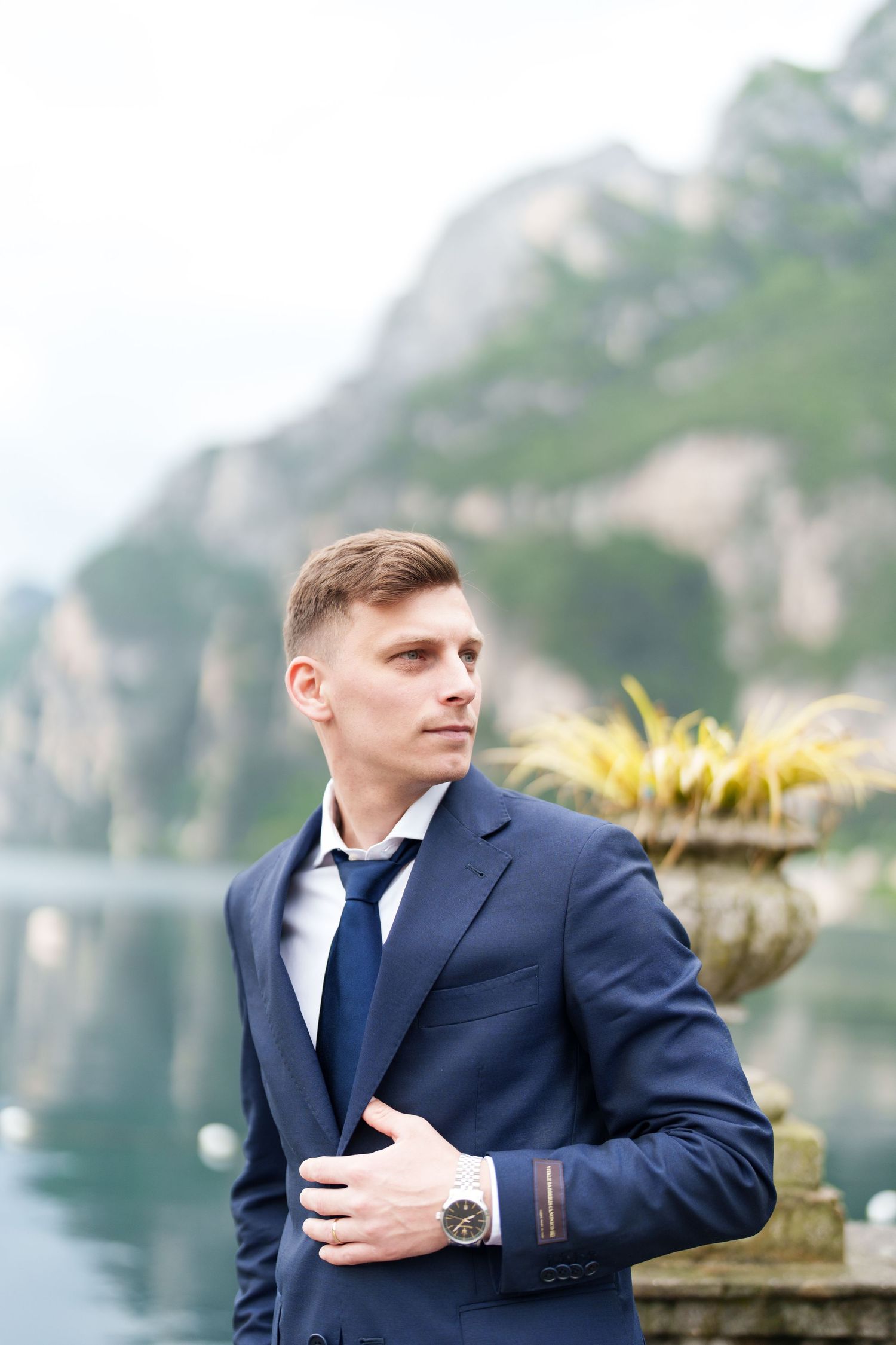 A groom in a navy suit poses thoughtfully against a scenic mountain lake backdrop with decorative stone elements.