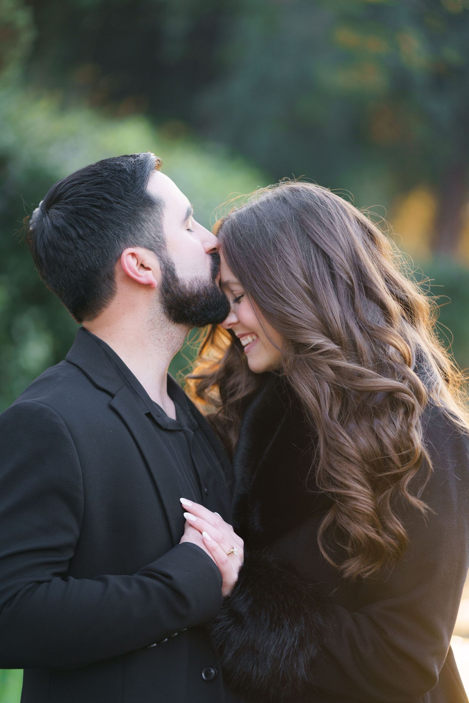 A romantic couple in black clothing share an intimate kiss outdoors with blurred natural background.